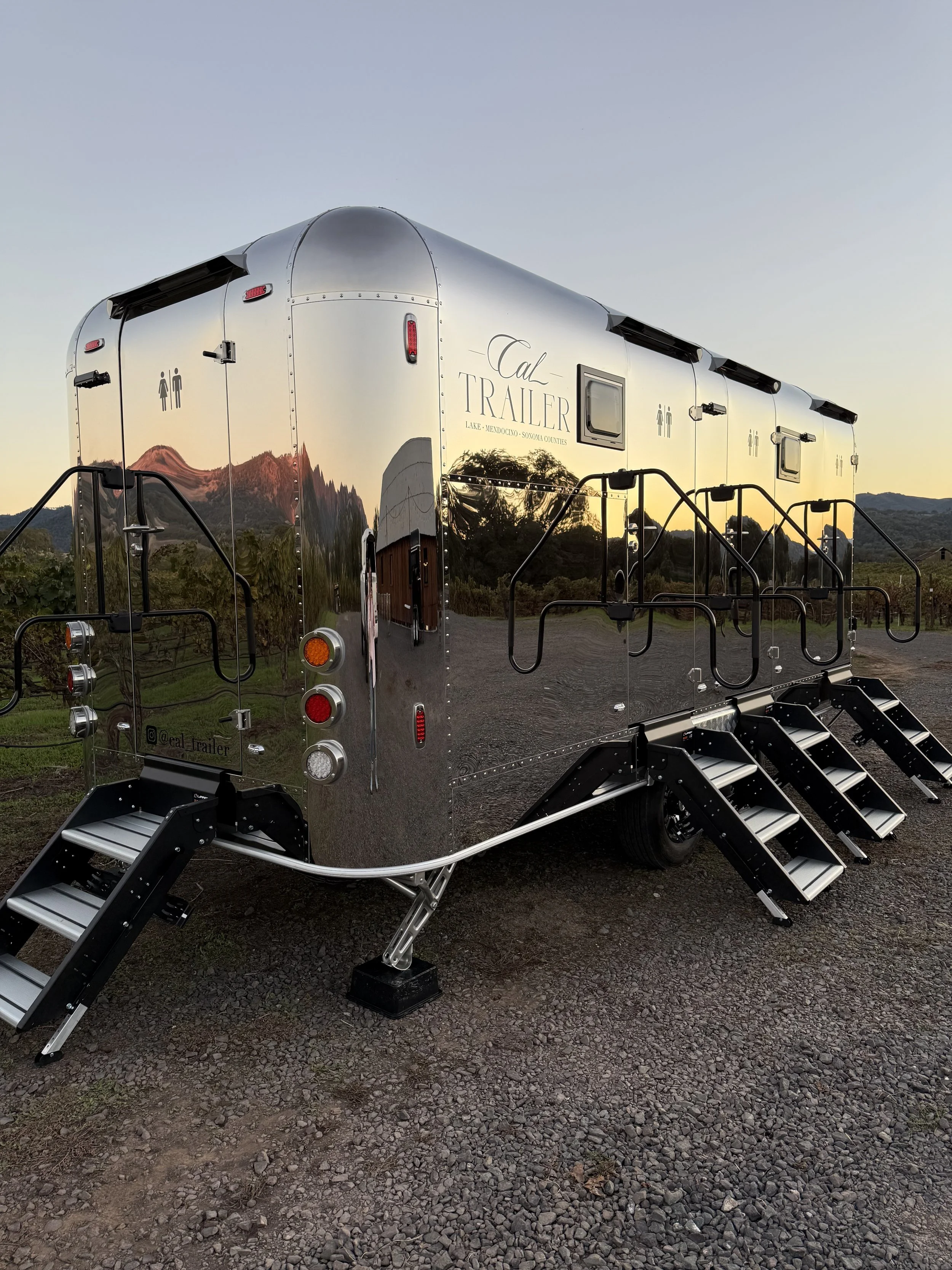 A shiny, metallic trailer with stairs leading up to the entrance, set outdoors with a scenic backdrop of hills and trees, and a sign that reads 'Cal Trailer'.