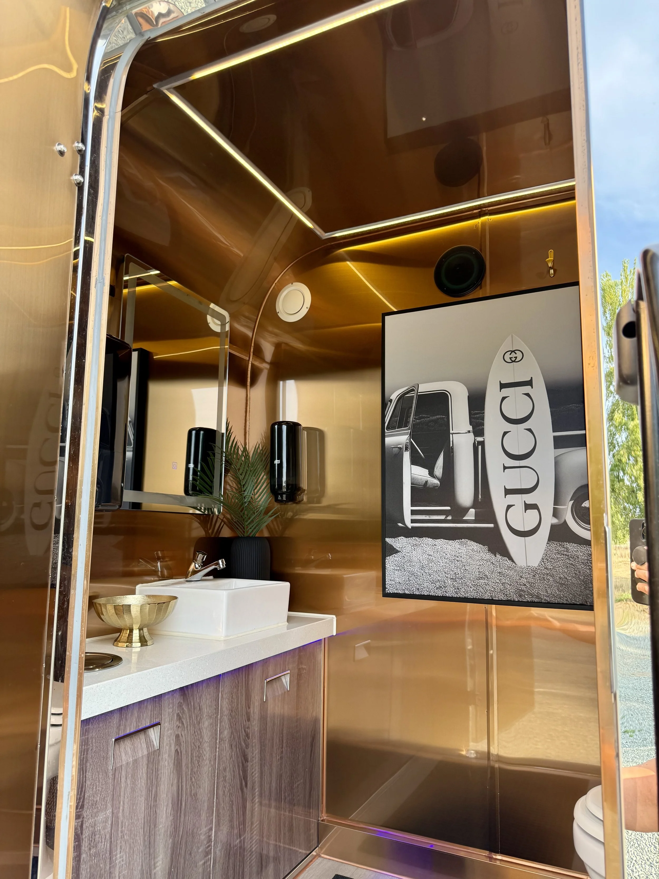 Interior of a luxury bathroom with gold-colored walls, a white sink, a gold bowl, black soap dispensers, a framed black and white photo of a truck with the GUC logo, and a potted plant.