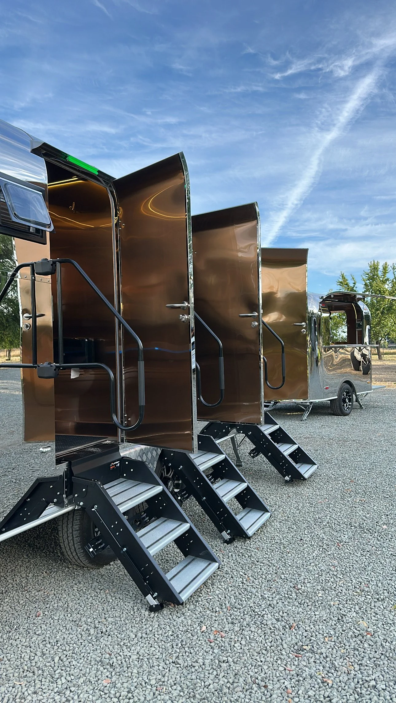 A mobile food truck with metallic brown doors, black stairs, and black rails, parked outdoors under a blue sky with wispy clouds.