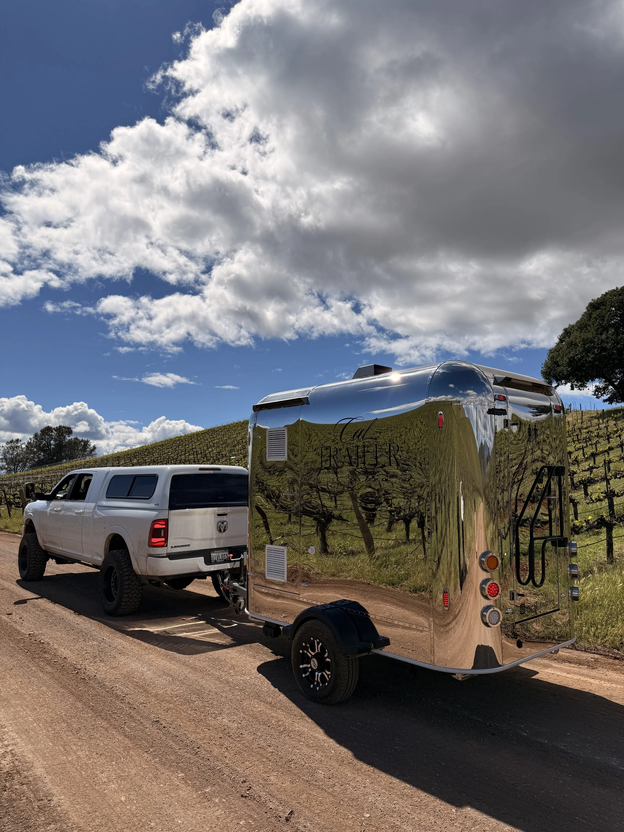 A white pickup truck parked on a dirt road next to a shiny silver trailer with the words Cal Trailers on it, beneath a sky filled with large fluffy clouds.