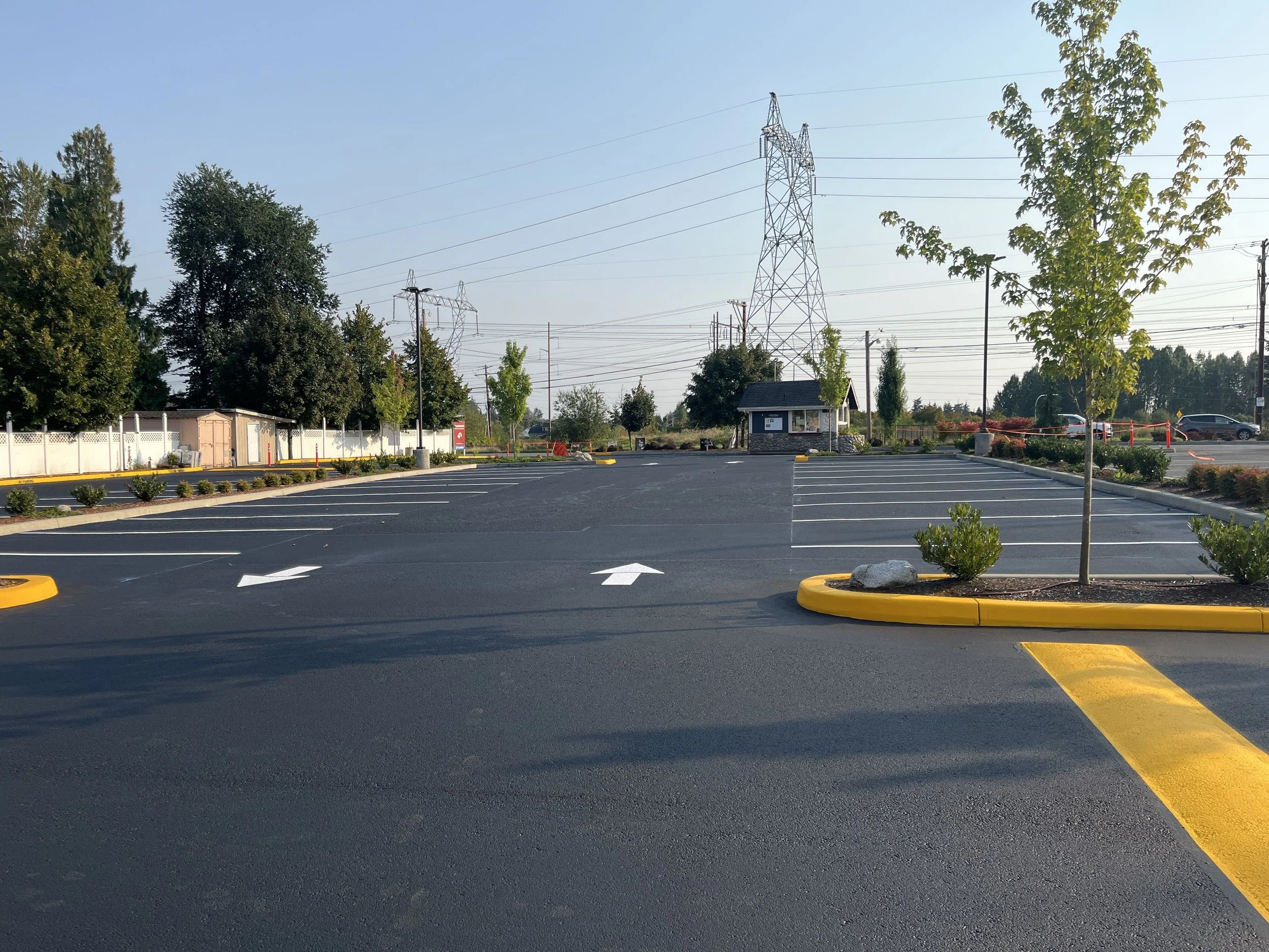 Empty parking lot with a few small trees, a white fence, and utility poles and power lines in the background on a clear day.