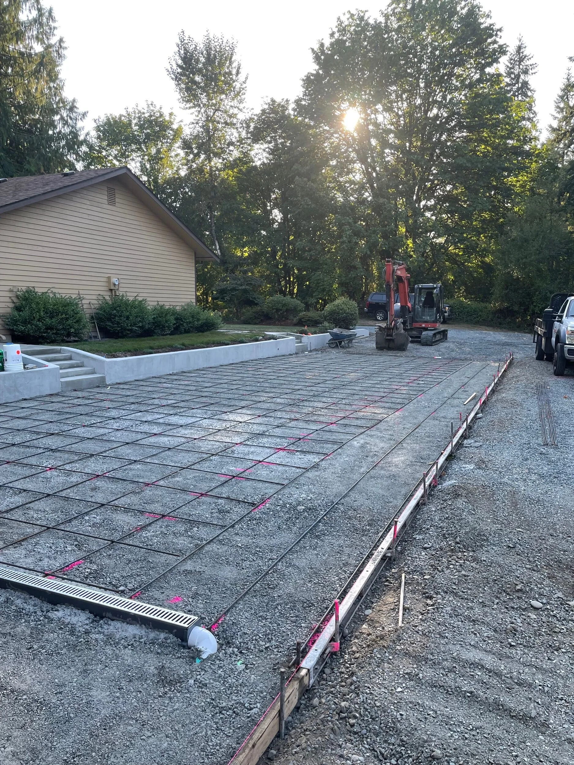 Construction site with concrete pouring and rebar grid, small excavator, parked vehicles, a house, trees, and the sun in the background.