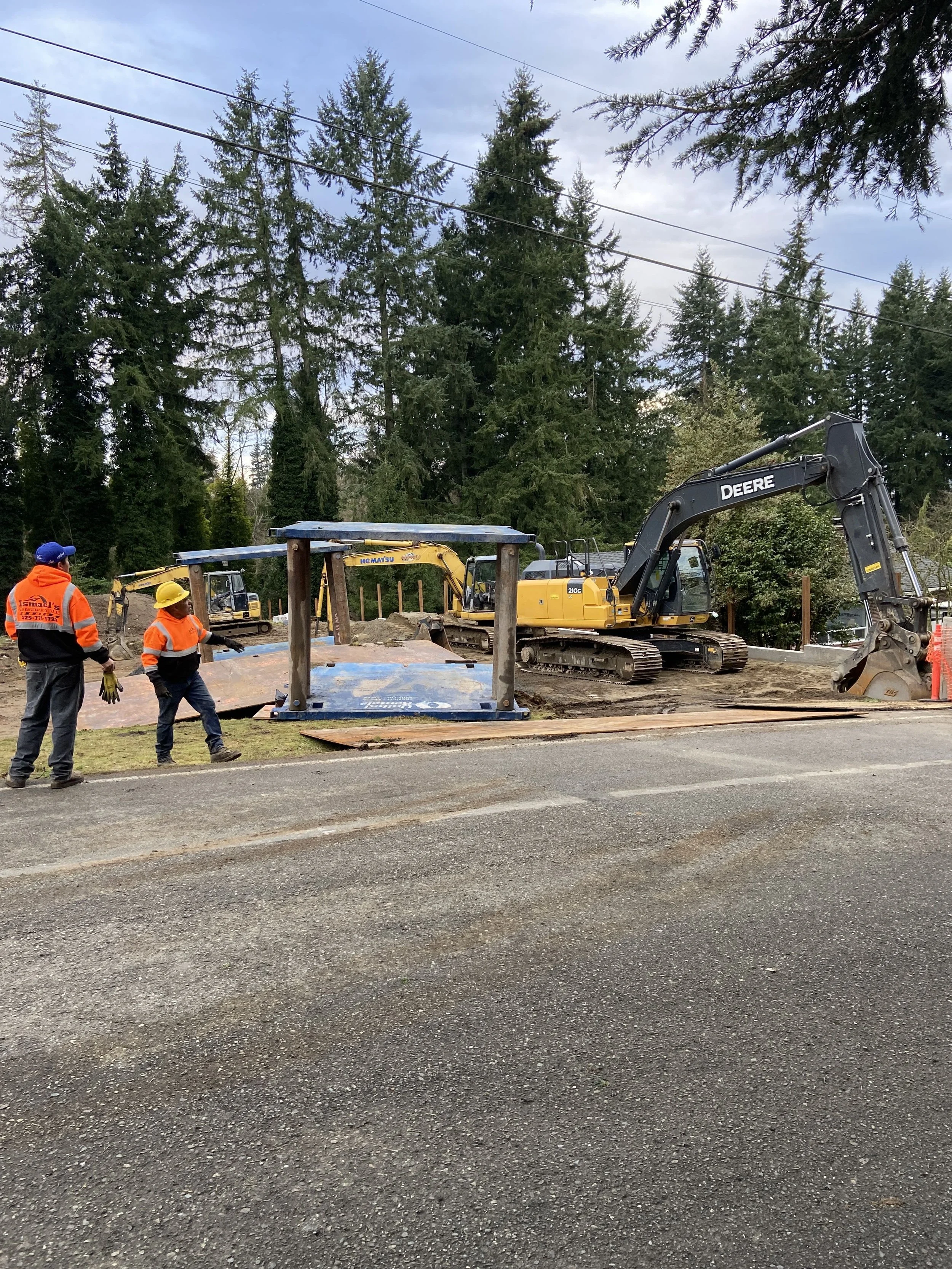 Construction workers in orange safety vests and yellow hard hats working on a road project with construction equipment, including an excavator and construction materials, in a forested area.