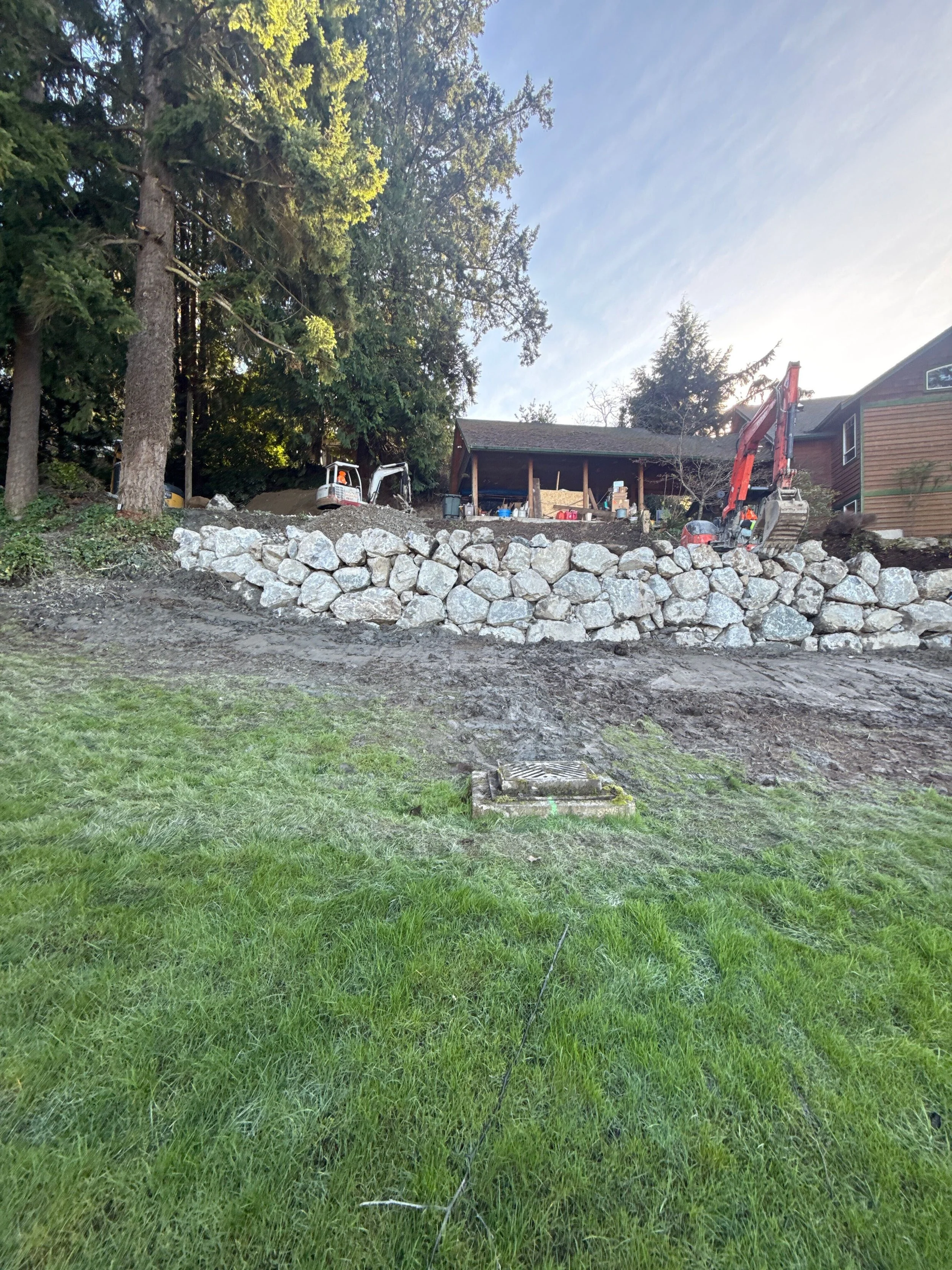 A backyard under construction showing a stone retaining wall, a grassy lawn in the foreground, a dense forest of tall trees, and a small building with construction equipment including a mini excavator, a construction worker, and supplies.