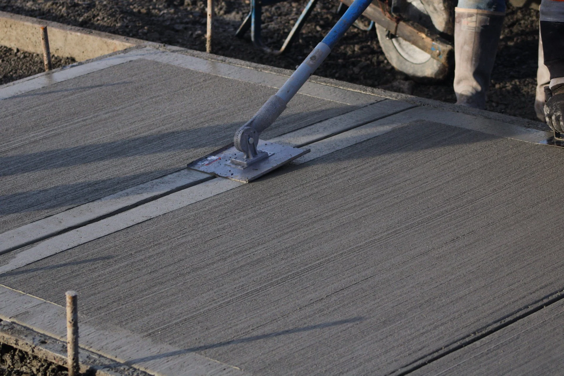 A person is using a hand trowel to smooth wet concrete on a newly poured sidewalk or driveway, with a wheelbarrow and construction background visible.