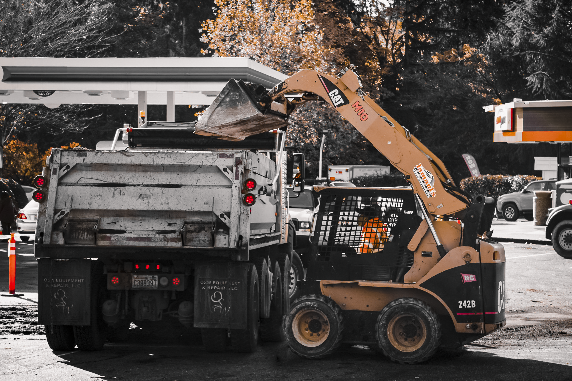 Construction worker operating heavy machinery in parking lot, with trucks and cars in the background, and a store canopy overhead.