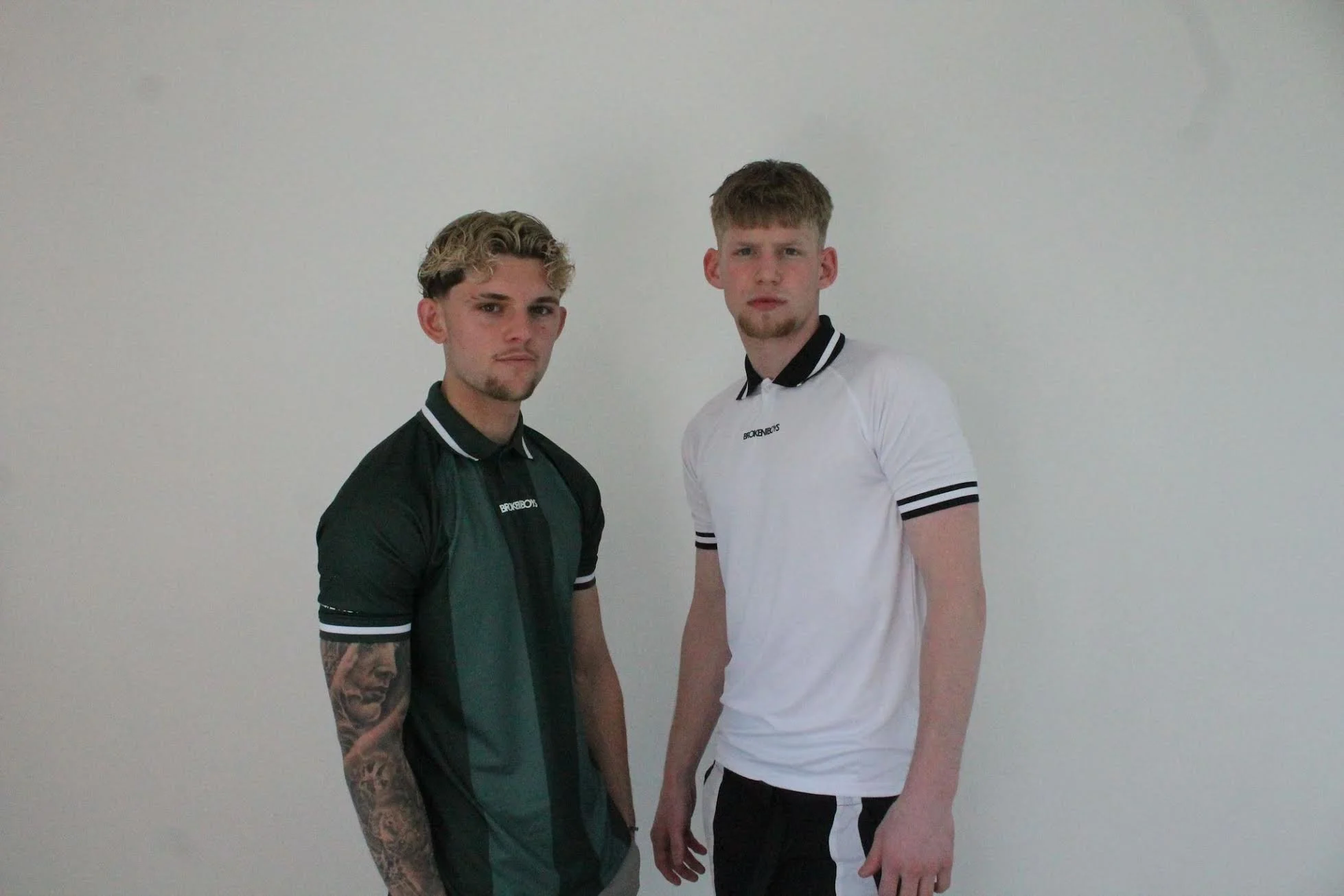 Two young men standing against a plain white wall, both wearing casual athletic clothing, looking at the camera with neutral expressions.