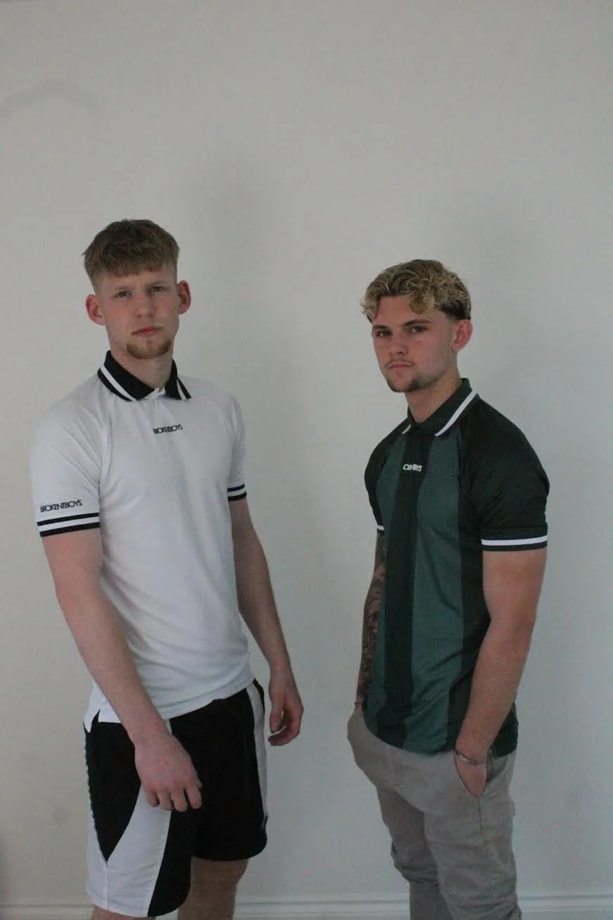 Two young men standing against a plain light-colored wall, both with serious expressions, wearing casual golf clothing called brokentboys