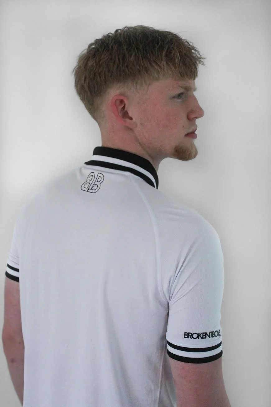 Side profile of a young man with red hair and a short beard wearing a white sports shirt with black stripes and logo, standing against a white background.