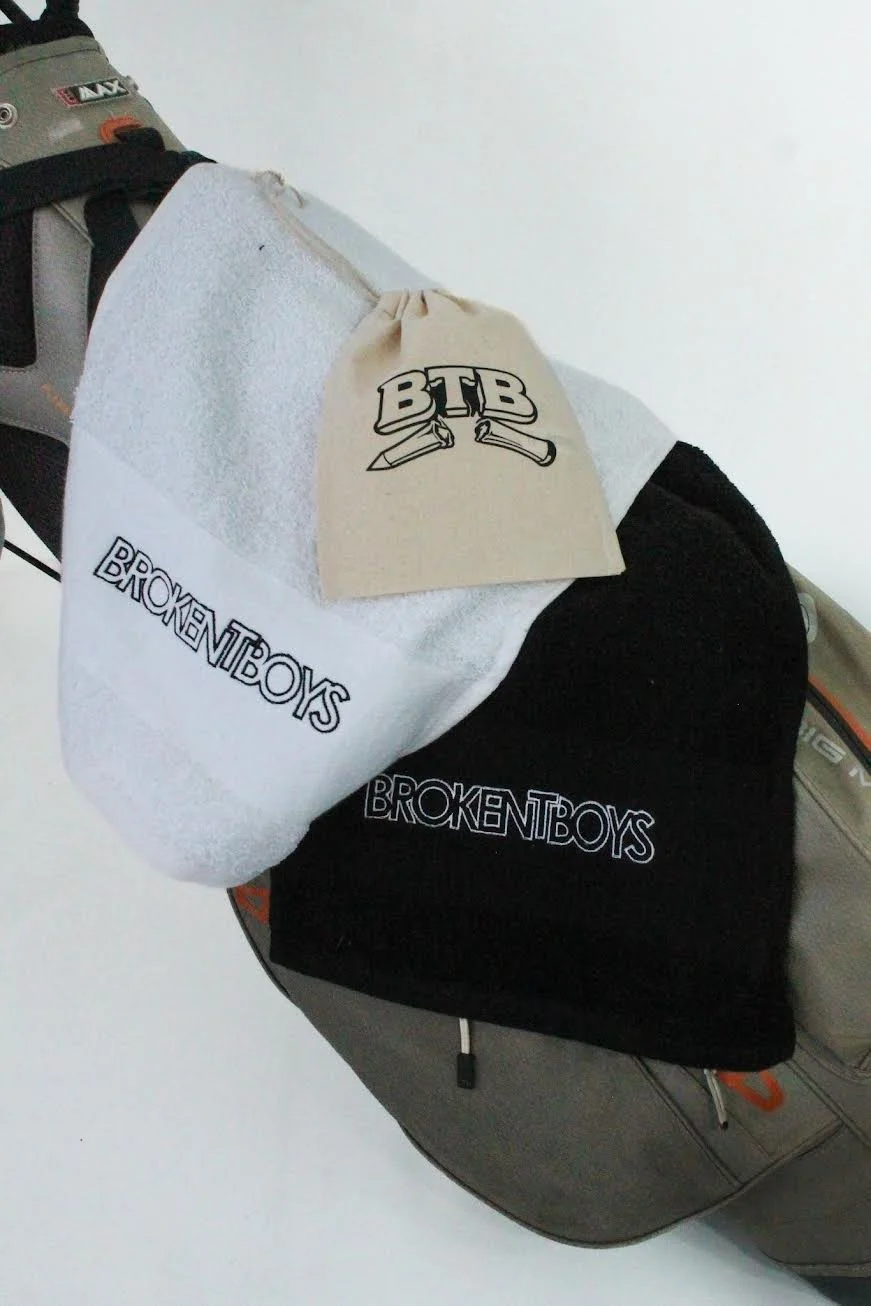 A golf bag with a white towel, a beige drawstring bag with the letters 'BTB' and two knives, and a black beanie with the text 'BROKENBOYS' embroidered on it.