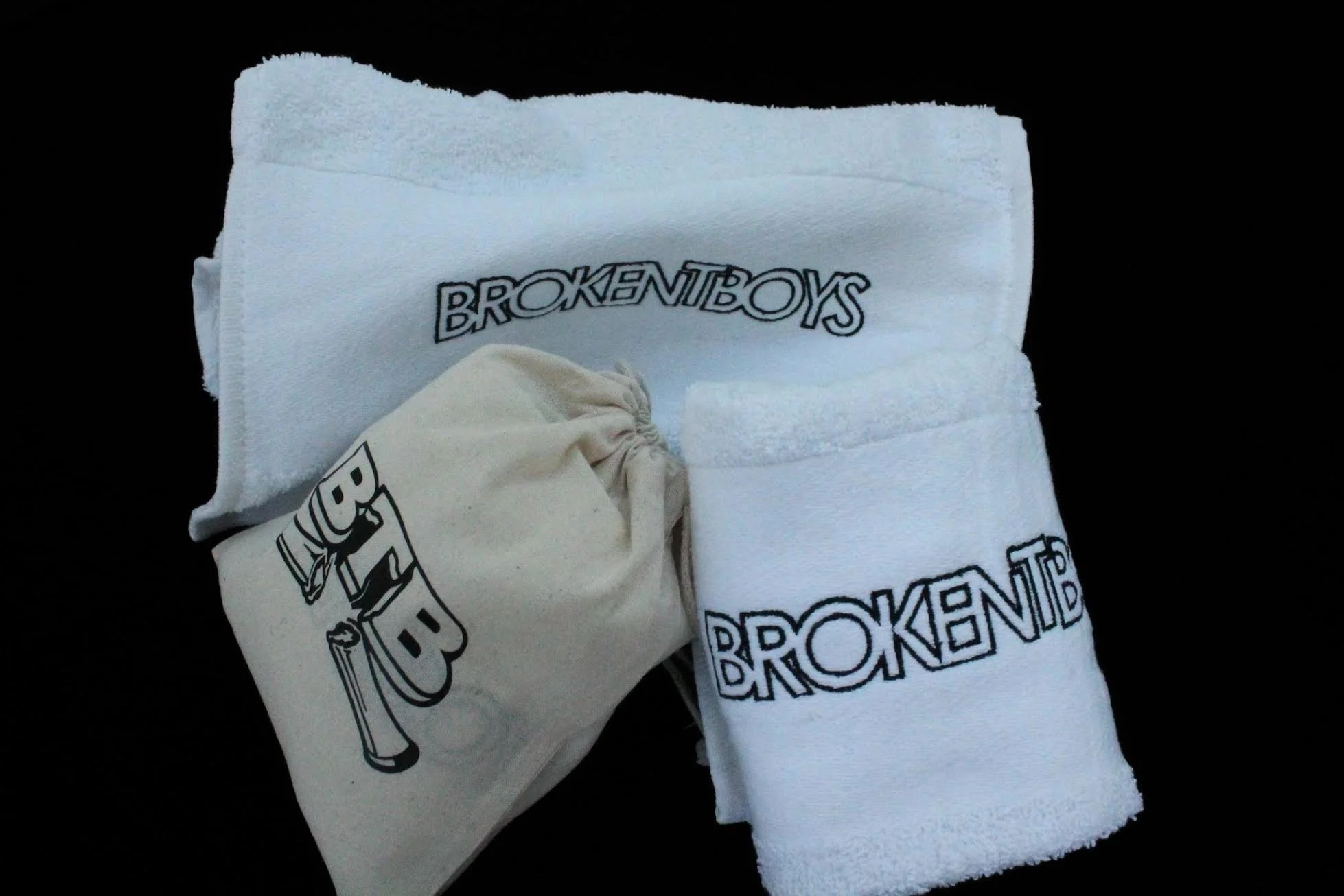 A white towel and a cloth bag with the words 'BrokentBoys' on them, placed on a black background.