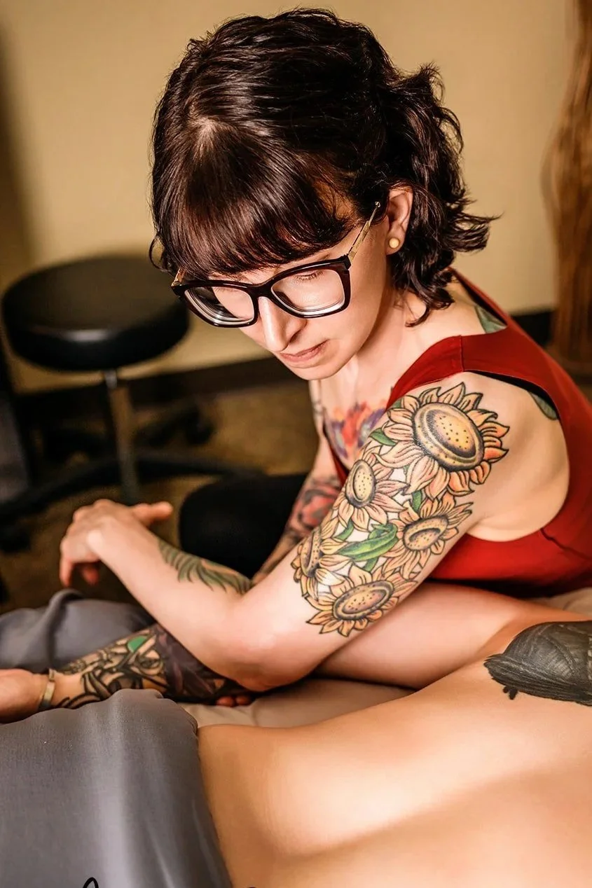 A woman with dark, short hair and glasses, wearing a red top, showing her colorful sunflower tattoos on her left arm and shoulder, is sitting on a bed or lounge.