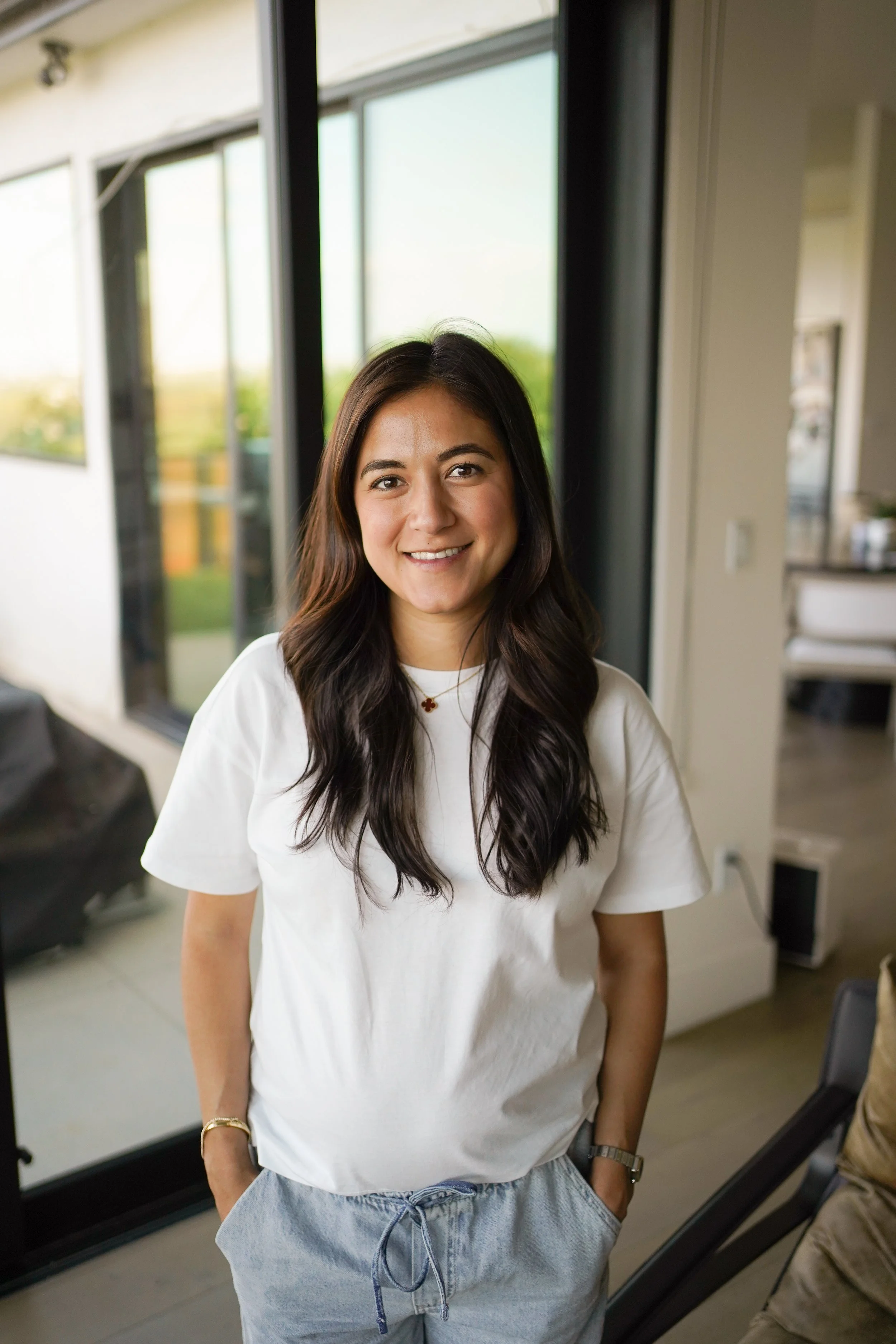 A young woman with long, dark hair, smiling, standing indoors near a large window with a green outdoor view. She is wearing a white T-shirt, light blue jeans, and accessories including a bracelet and a necklace with a cross pendant.