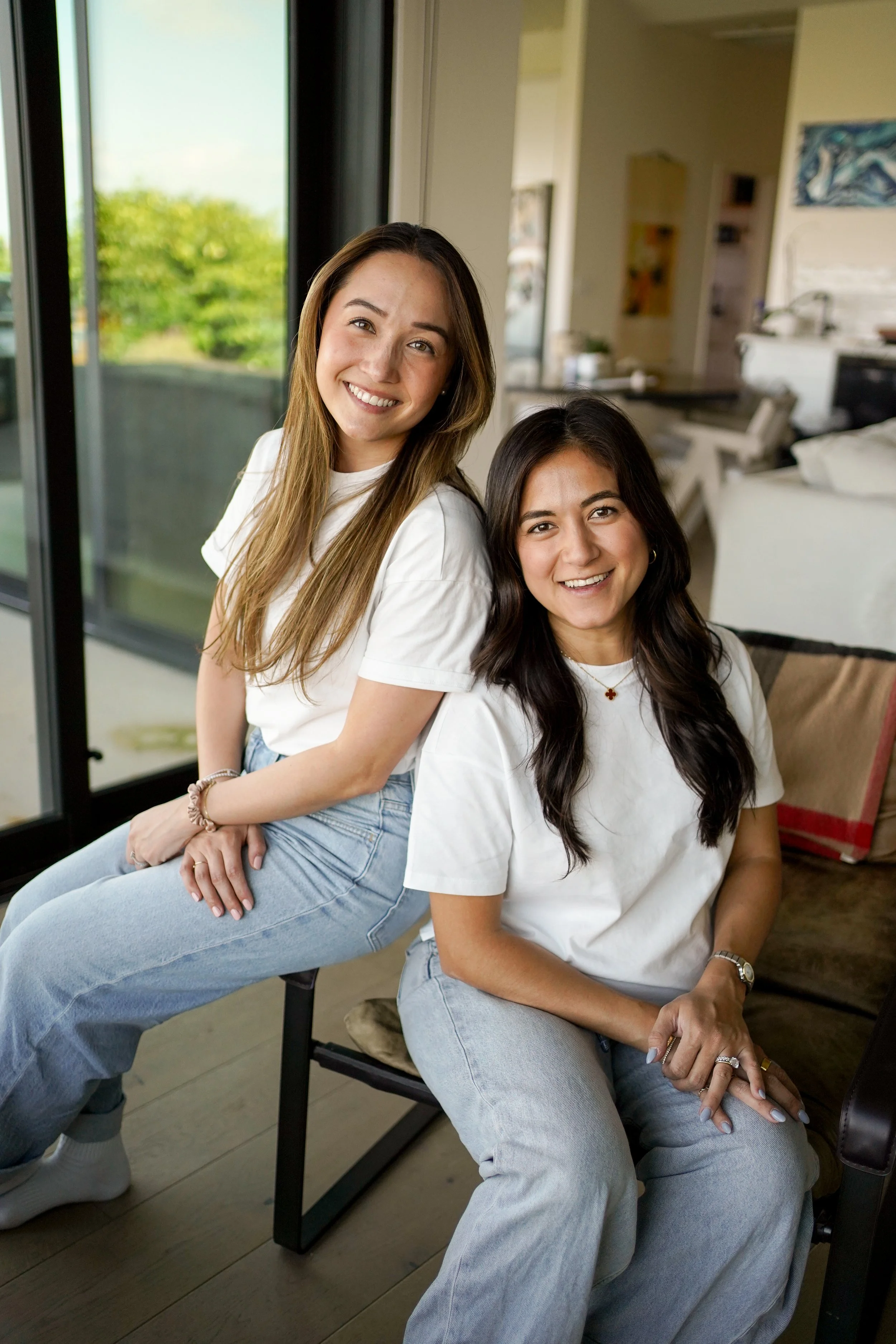 Two young women sitting in a cozy indoor space near large glass doors, smiling and looking at the camera. One has long brown hair, and the other has long dark hair. Both wear white t-shirts and light-colored jeans.