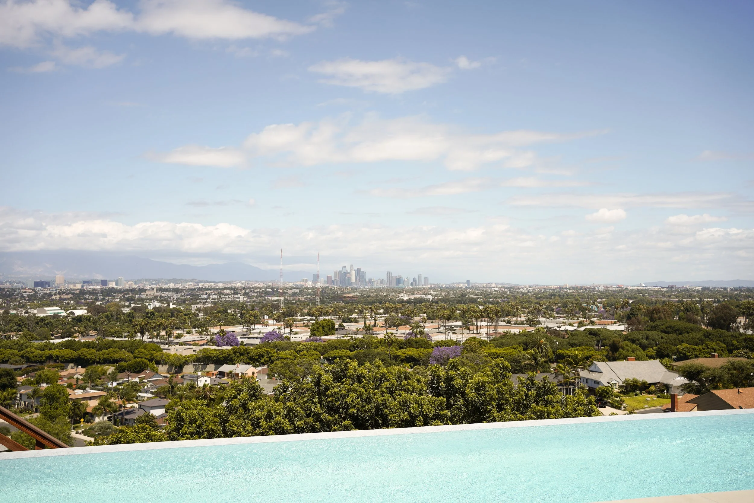 City skyline with buildings, trees, and mountains in the background, viewed from a rooftop pool with a white edge and turquoise water in the foreground.