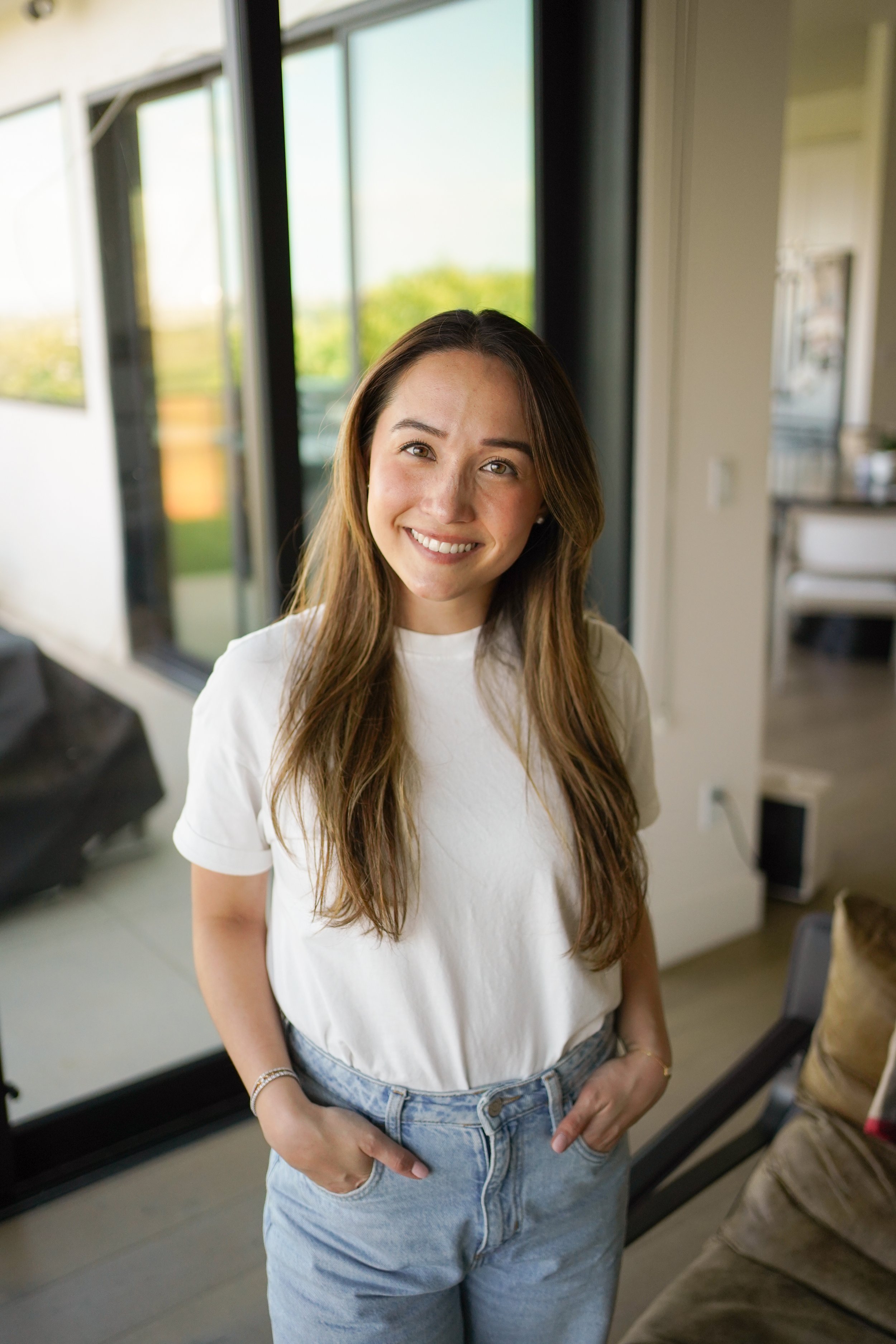 A young woman with long brown hair, wearing a white t-shirt and jeans, stands indoors near large glass windows with greenery outside, smiling at the camera.
