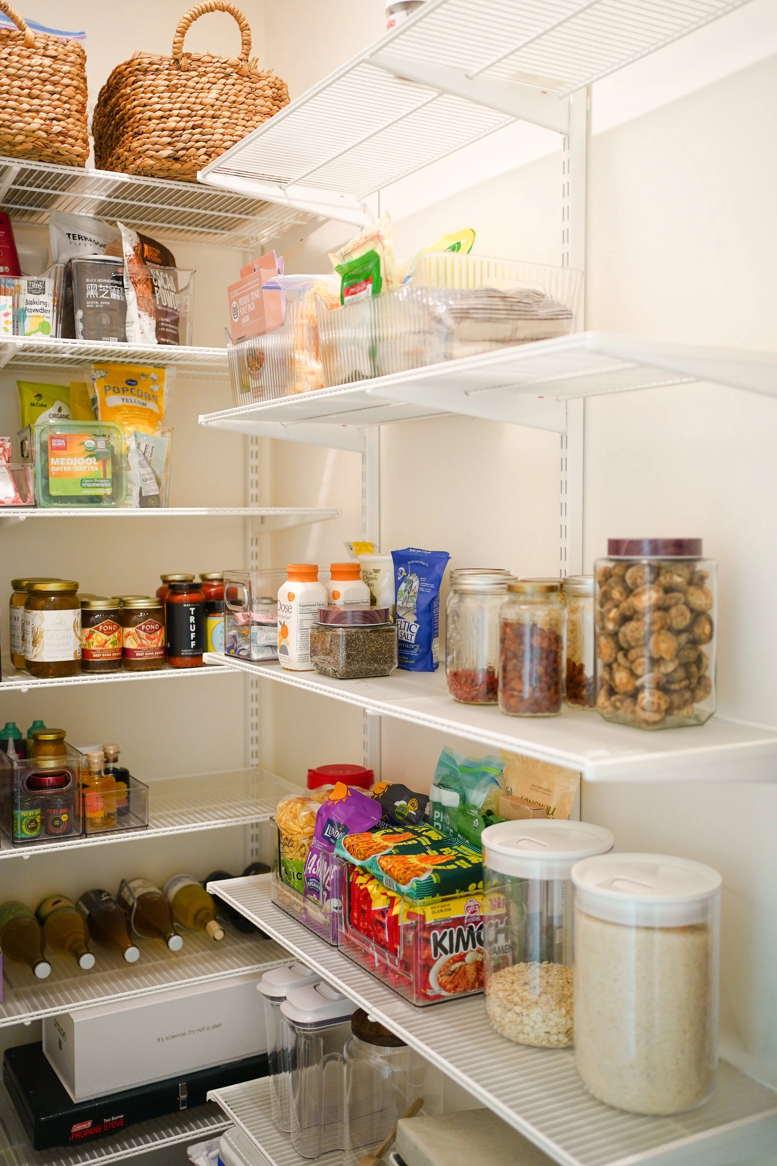 A pantry with white wire shelves filled with various food containers and jars, including dried goods, canned items, snacks, and bottles. Organized by Tori and Ash from Curated by AT.