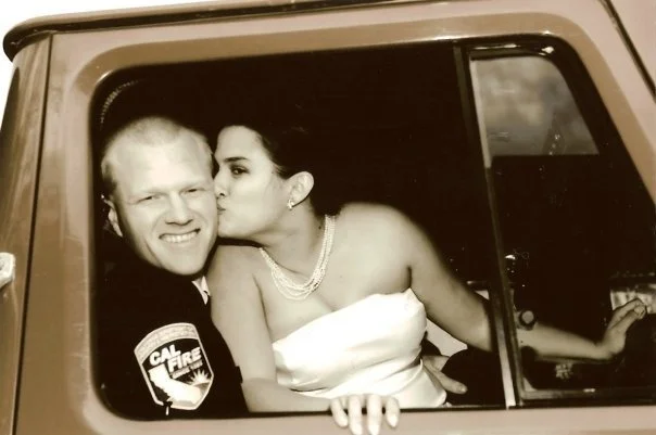 A woman in a wedding gown kissing a smiling male firefighter in a firetruck window.