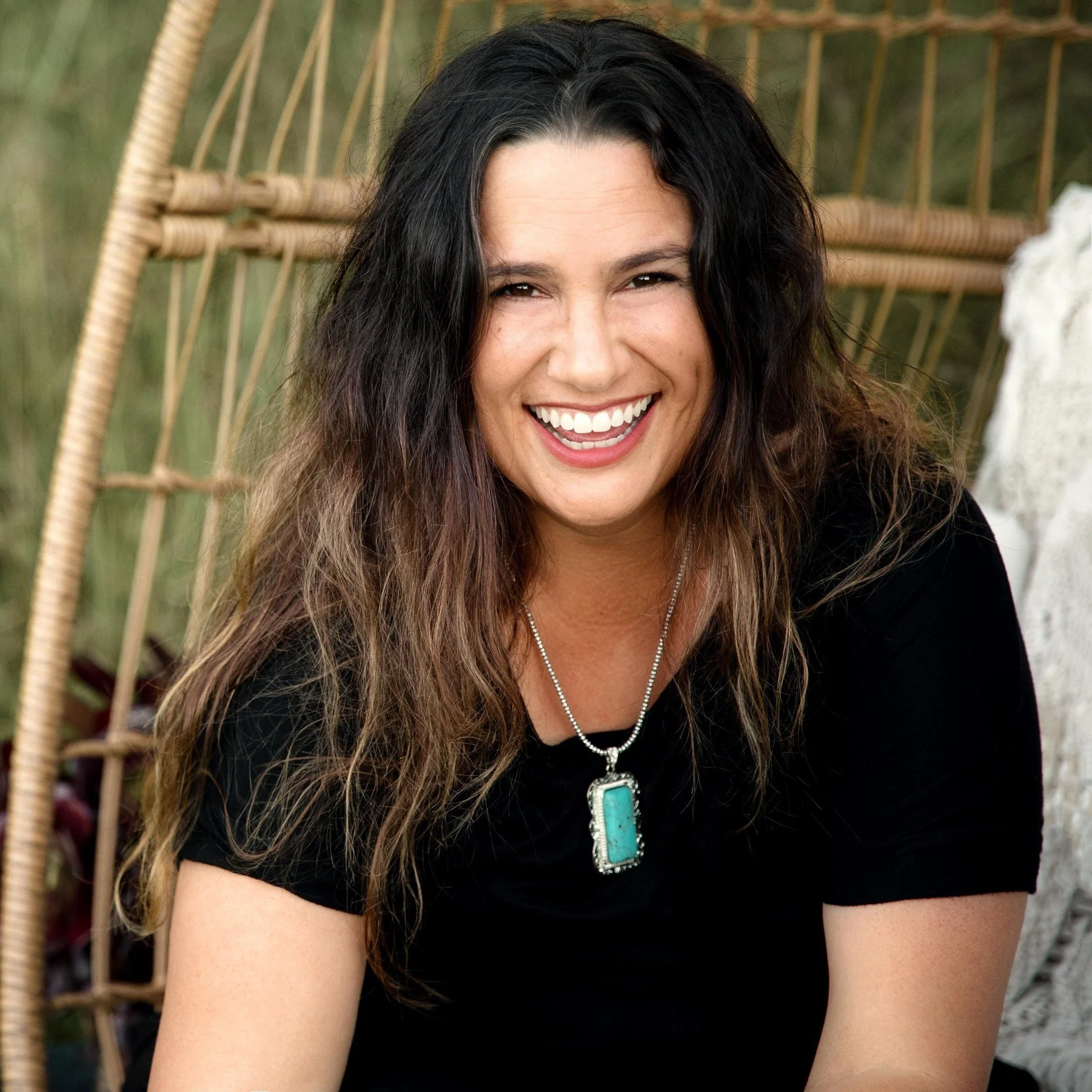 A woman - Chelsi McFadden- with long, curly dark hair smiling, wearing a turquoise pendant necklace and a black top, sitting outdoors with a wicker chair and greenery in the background.