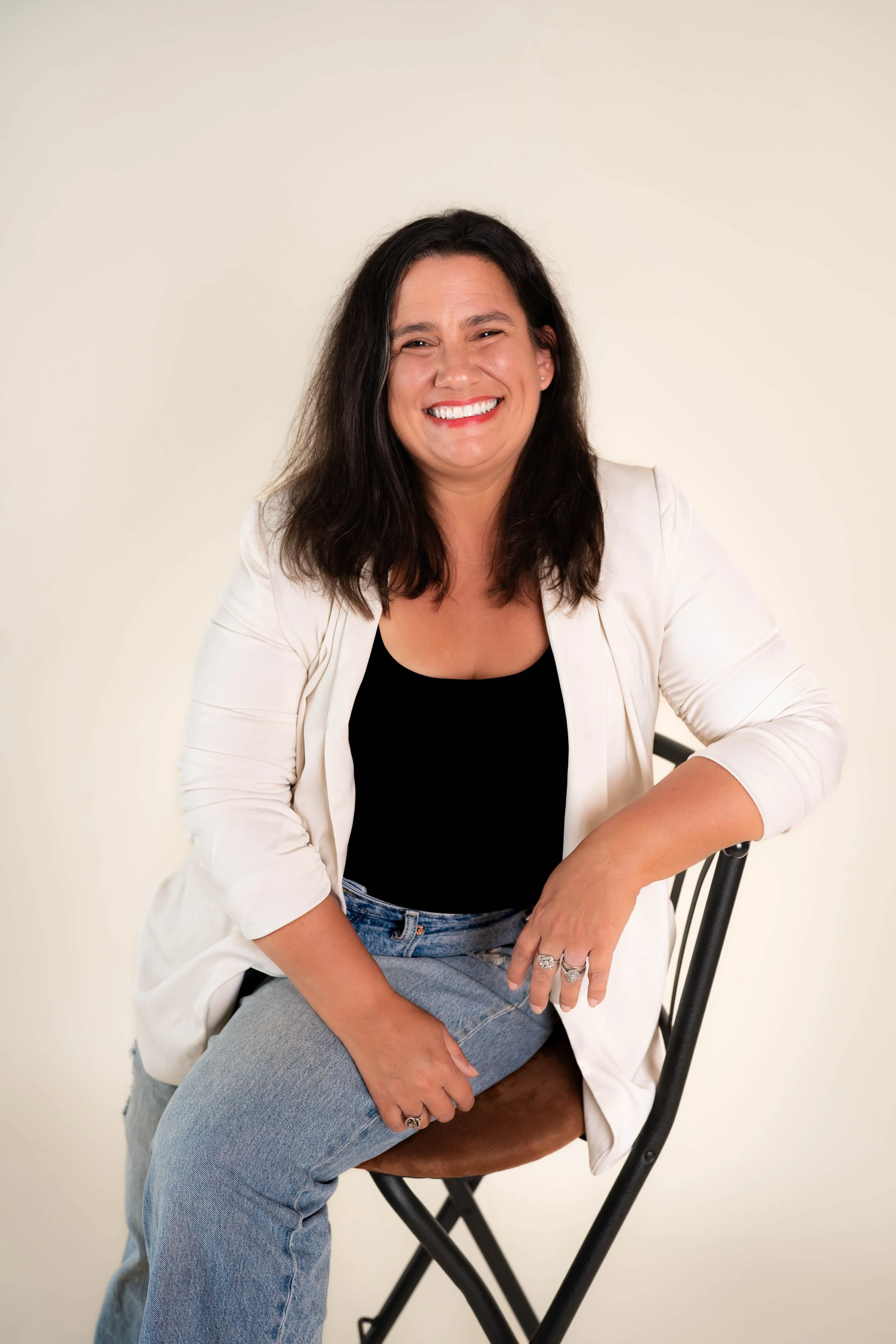 A woman - Chelsi McFadden - with long dark hair sitting on a black chair against a plain white wall, wearing a black top, white blazer, and light blue jeans, smiling directly at the camera.