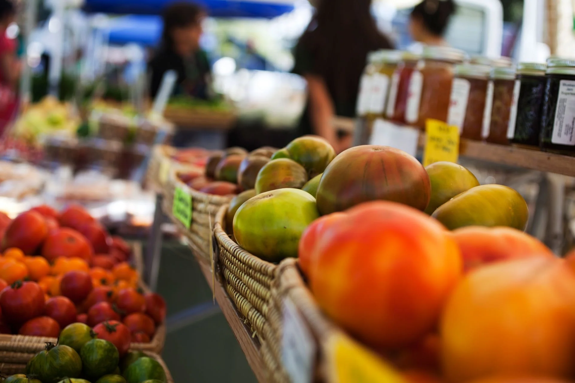 Basket of heirloom tomatoes at an outdoor farmers market with vendors and other produce in the background.
