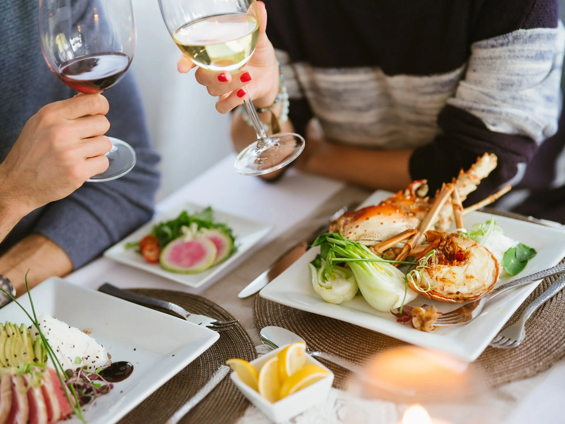 People holding glasses of white and red wine at a dinner table with various seafood and salad dishes.