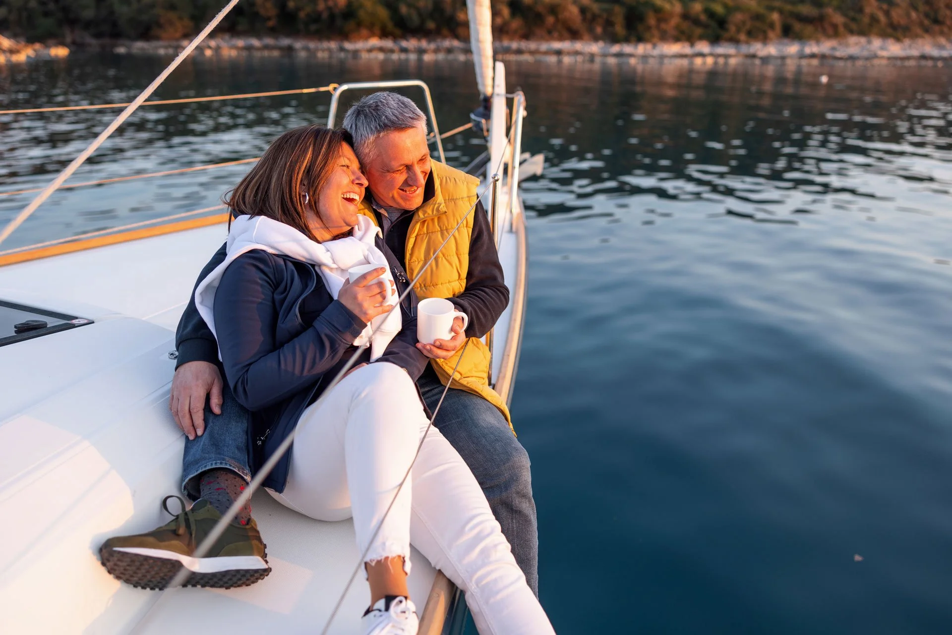 A couple sitting on a boat, laughing and holding cups of coffee, enjoying a scenic view of the water during sunset.