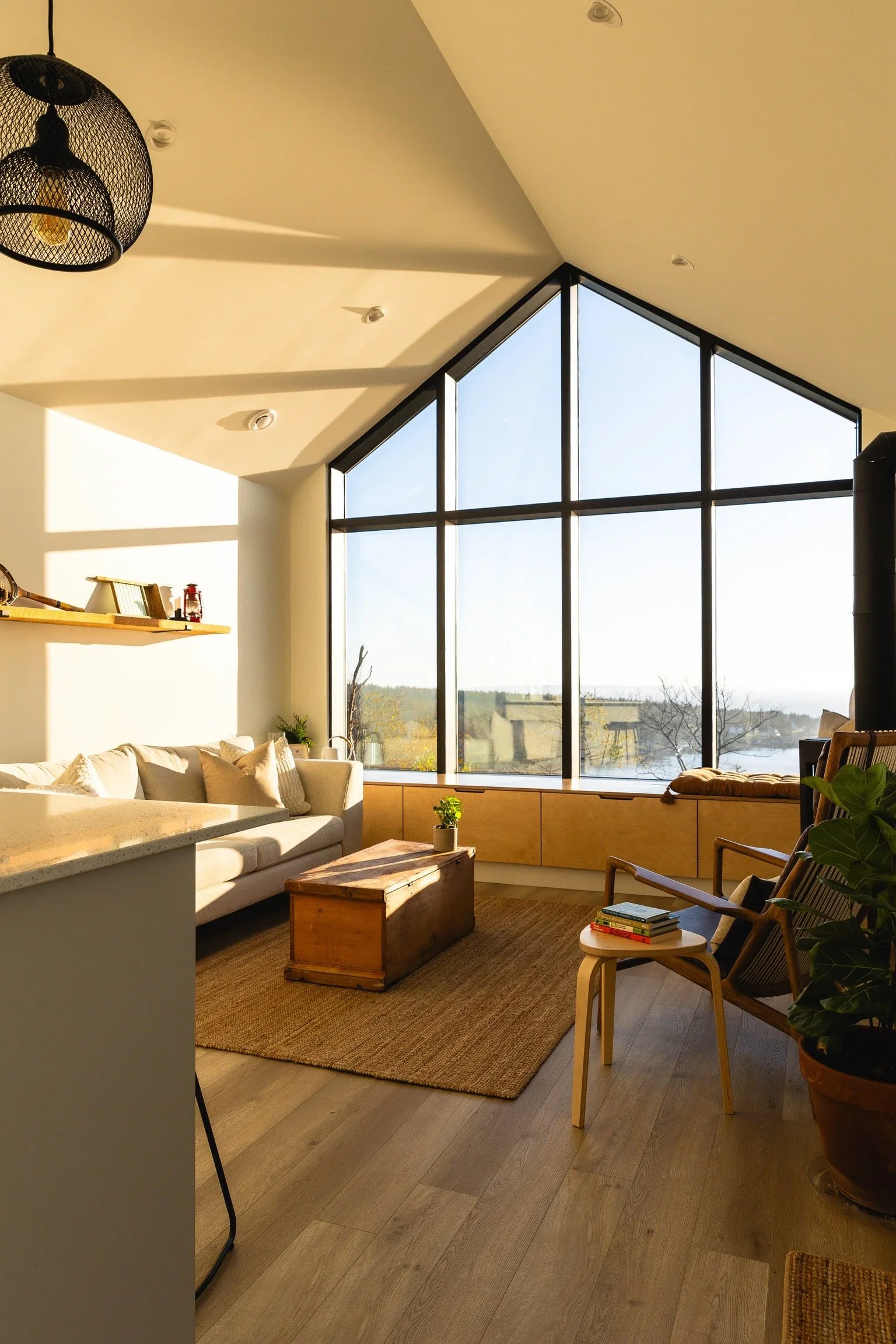 Living room with large triangular window, beige sofa, wooden coffee table, chairs, potted plant, shelf with decor, and natural light.