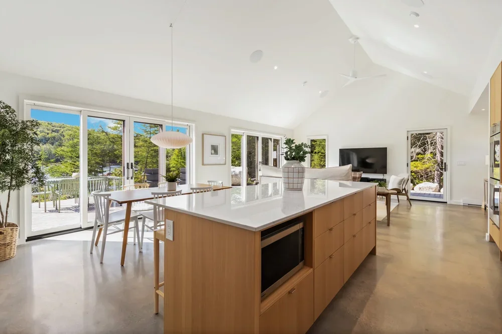 Open-concept kitchen and living room with large sliding glass doors leading to an outdoor deck, wooden dining table with white chairs, and a wall-mounted TV.