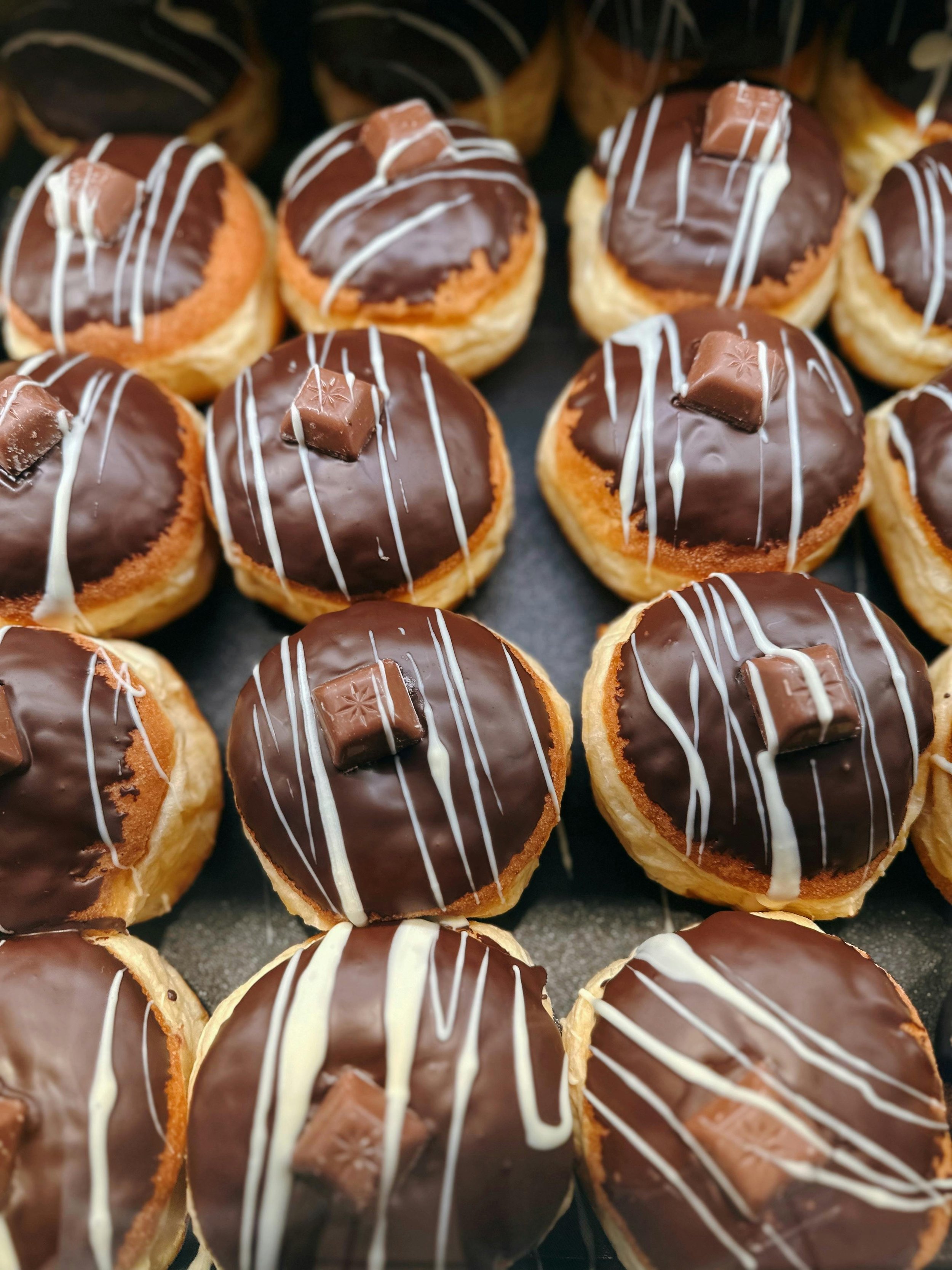 Close-up of multiple chocolate-glazed donuts decorated with white icing drizzles and topped with small chocolate squares on a tray.