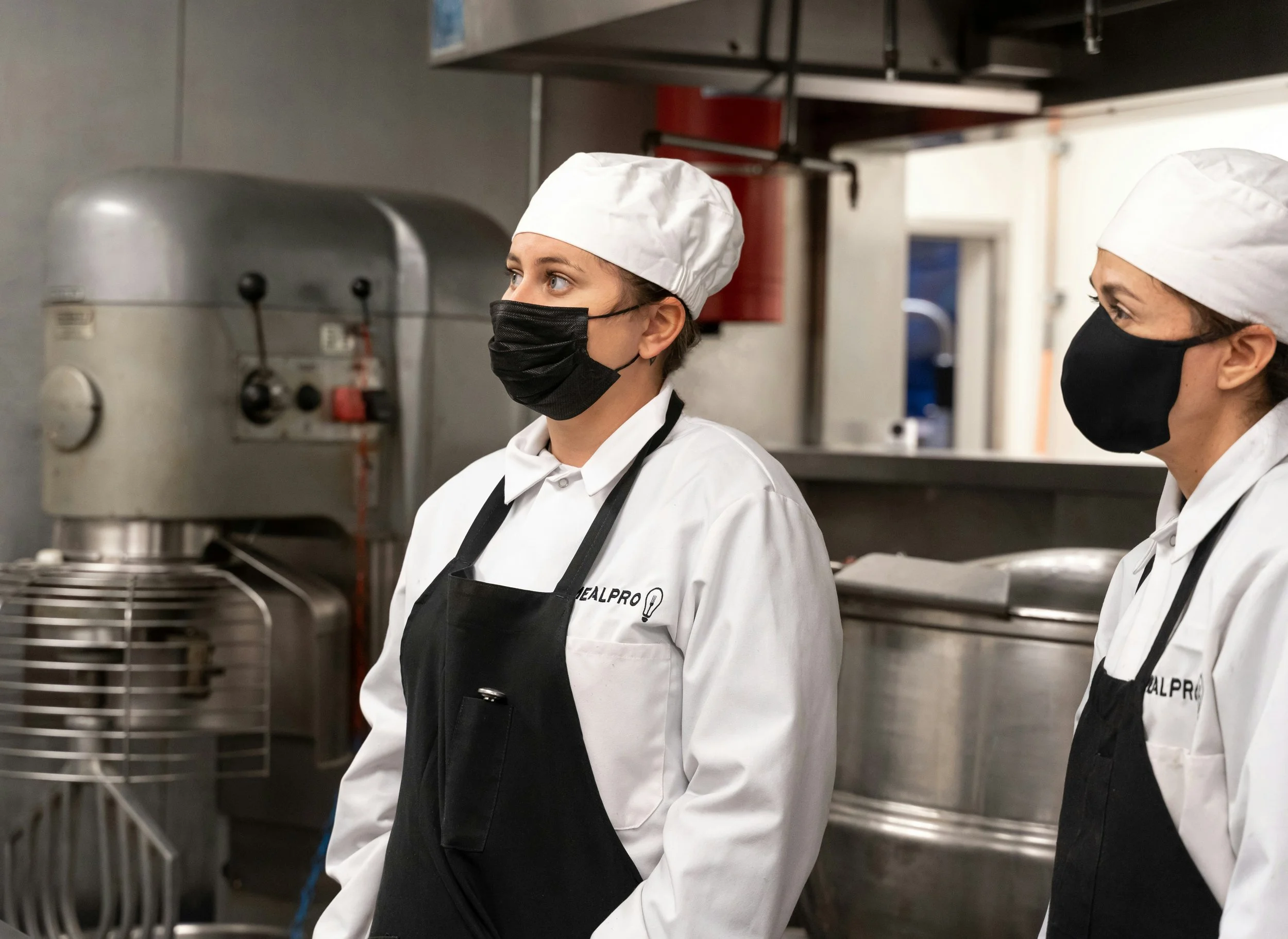 Two cooks in chef uniform and face masks working in a commercial kitchen with industrial kitchen equipment.
