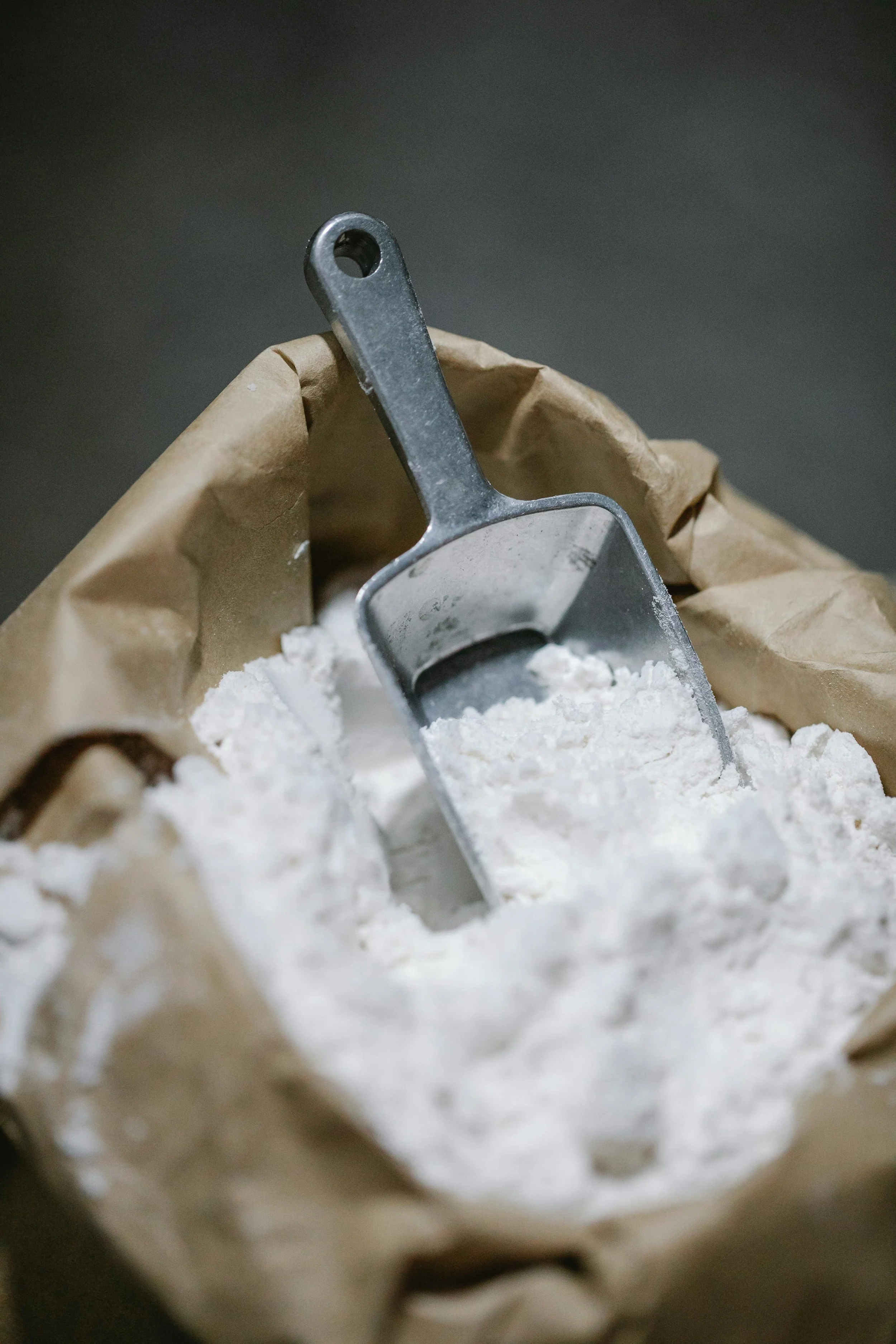 A metal measuring scoop is partially buried in white powder, within a crumpled brown paper bag.