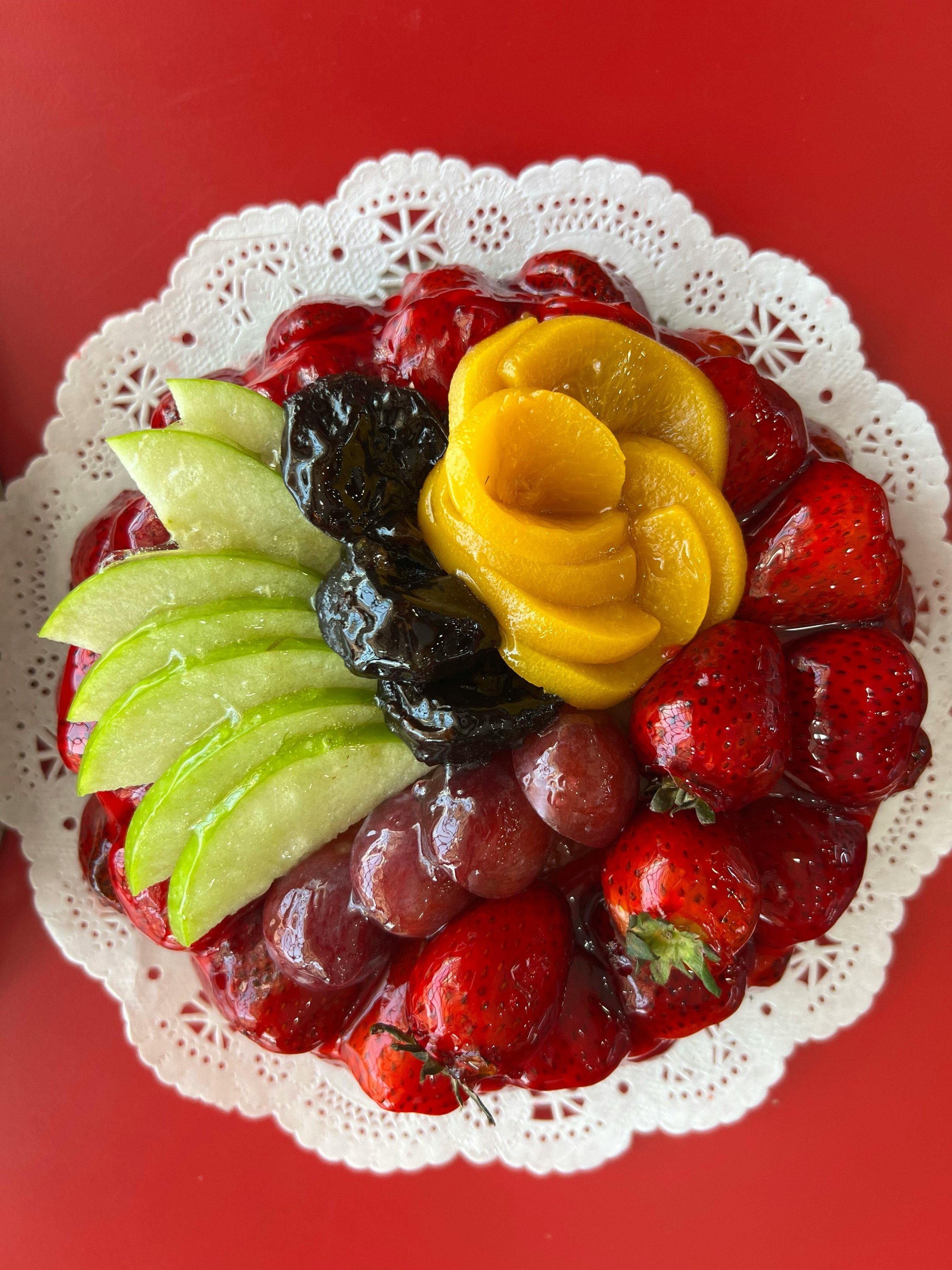 A plate of assorted fresh fruits including strawberries, green apple slices, blackberries, and yellow peach wedges on a decorative white doily placed on a red surface.