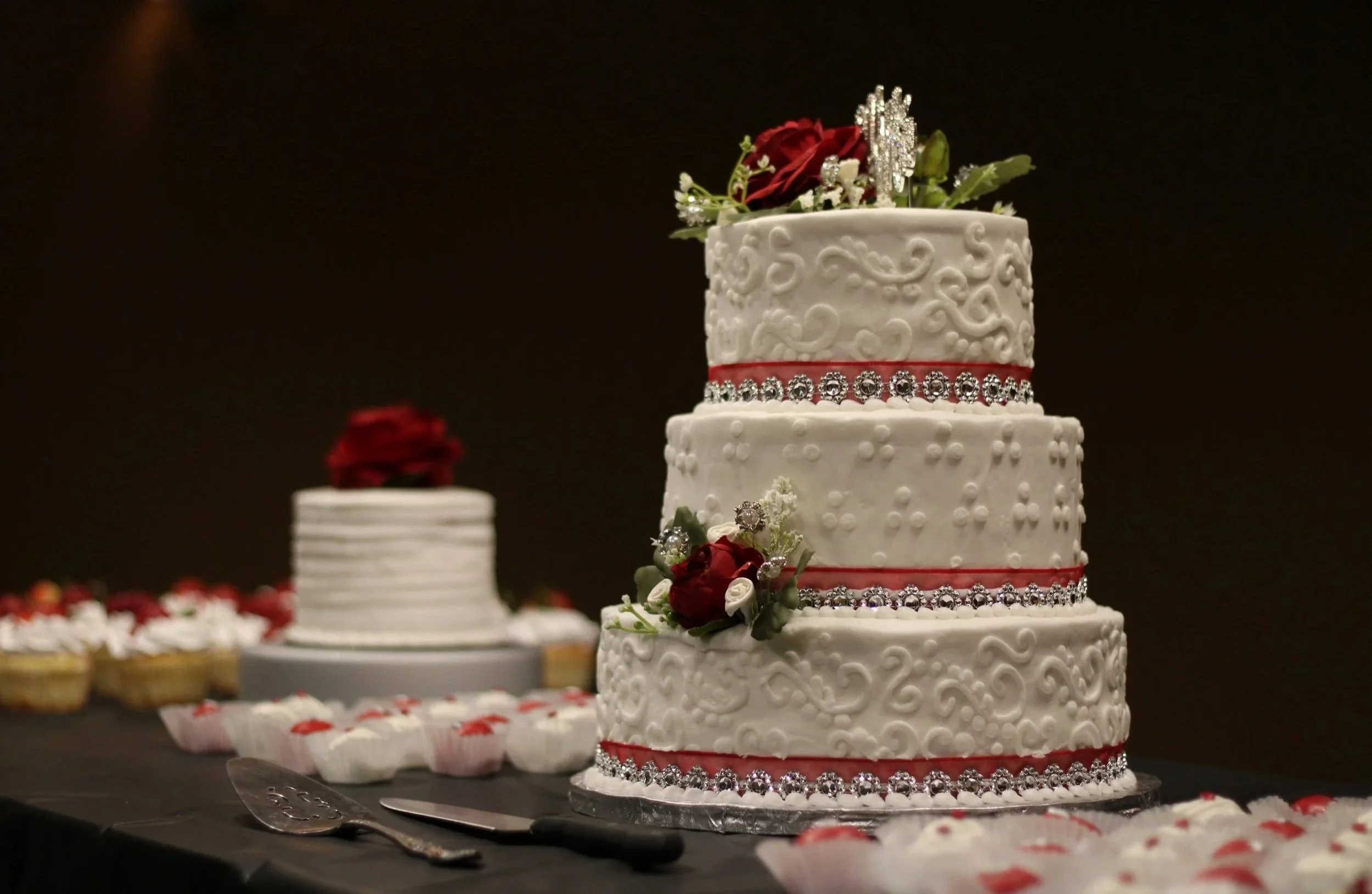 A decorative white wedding cake with intricate swirl patterns, red accents, and rhinestone embellishments, topped with red and white flowers and a silver tiara, set on a table with other desserts and red rose petals.