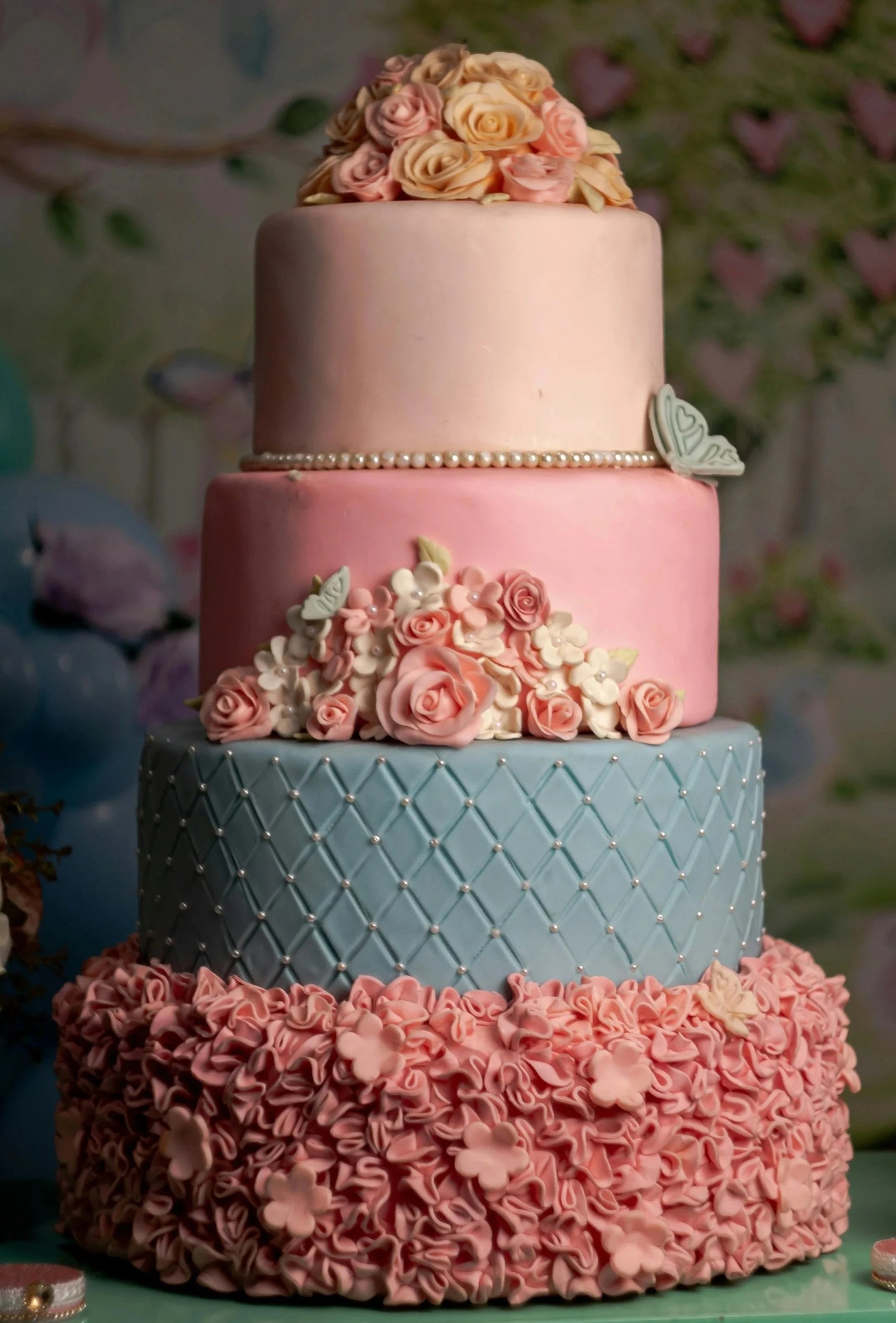 A multi-tiered wedding cake with pink and blue frosting, adorned with pink and cream flowers, pearls, and decorative butterflies.
