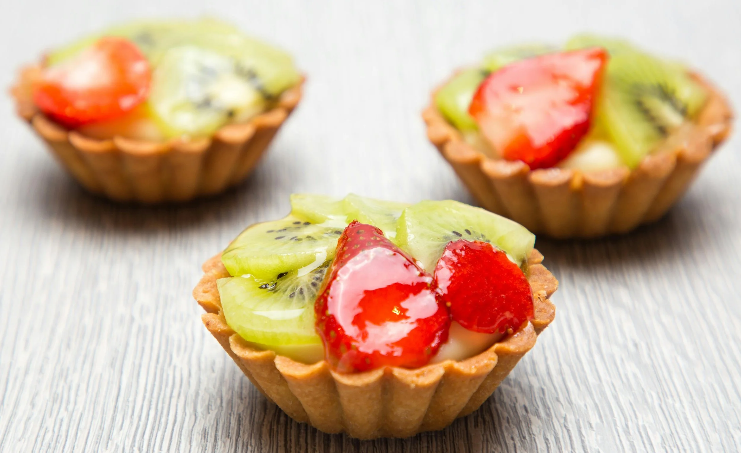 Three small tartlet cups filled with fresh strawberry, kiwi, and cream, placed on a light wooden surface.