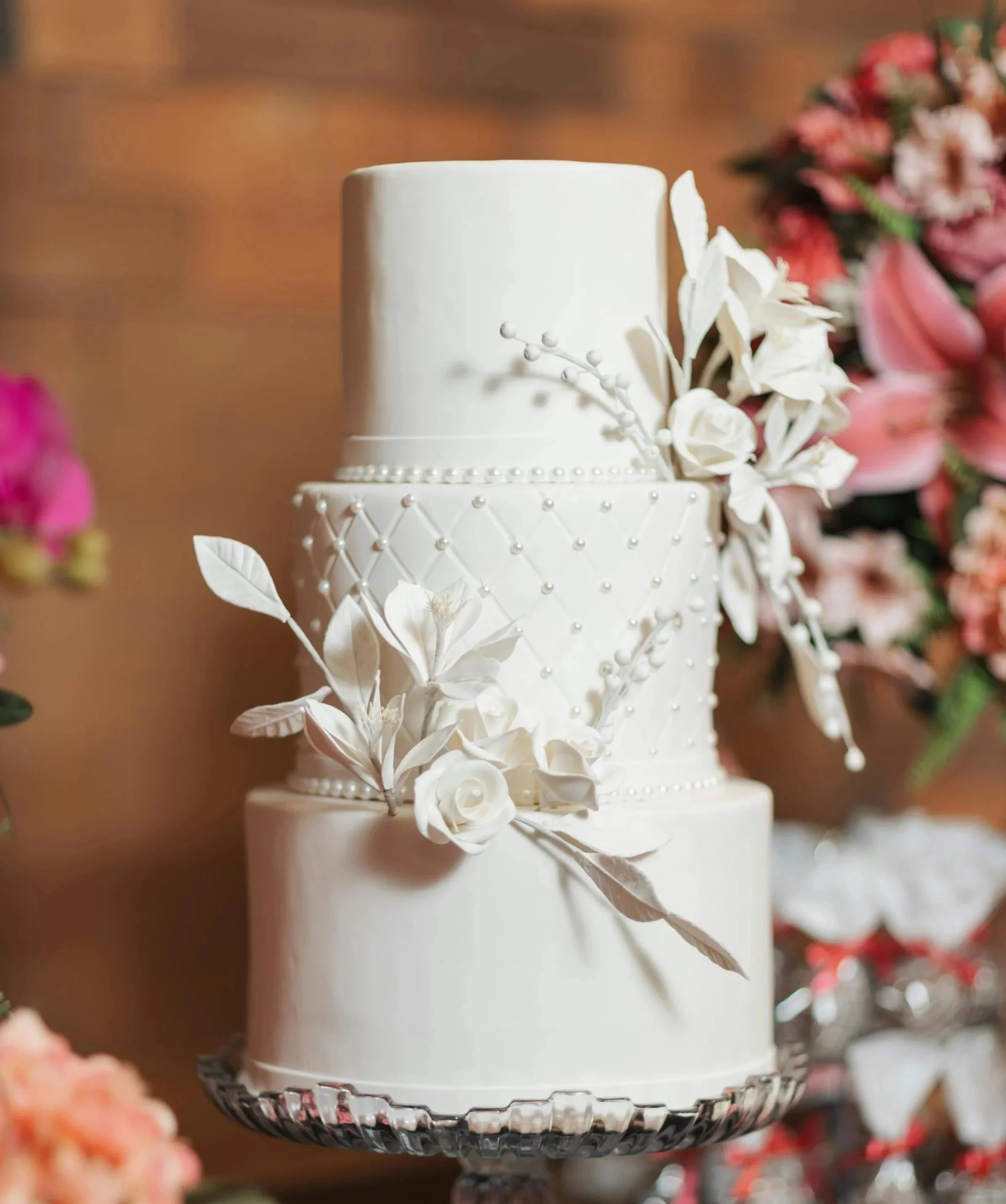 A white, multi-tiered wedding cake decorated with white sugar flowers and pearls on a silver cake stand.