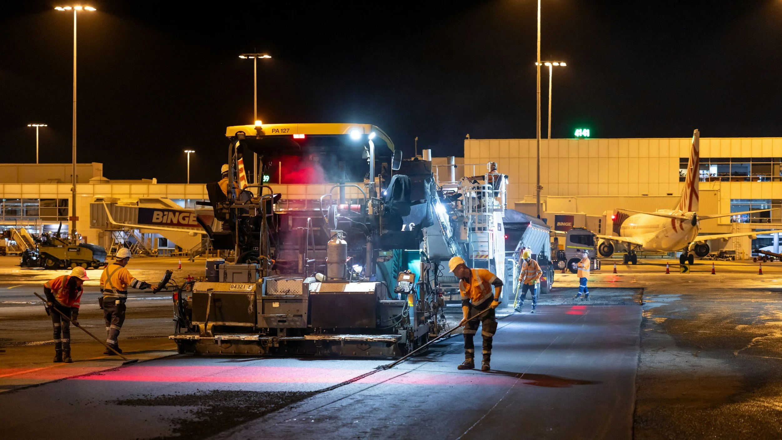 Nighttime scene at an airport runway where construction workers are laying down fresh asphalt. A large paving machine is in the center, with workers guiding and monitoring the process. In the background, an airplane is parked at the gate, connected to a jet bridge, with bright airport lights illuminating the scene.