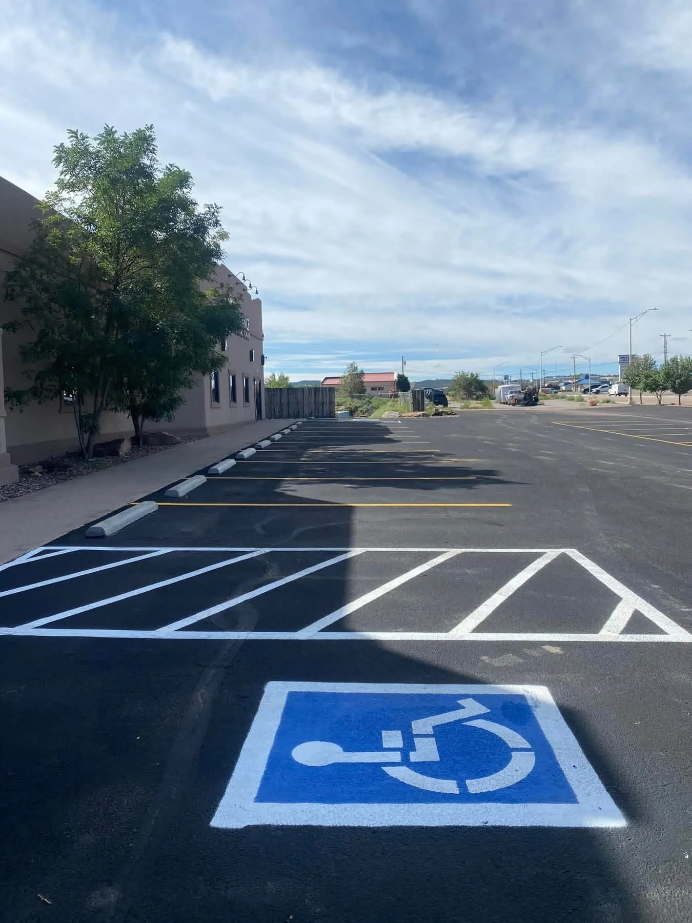 Empty parking lot with designated handicap parking spot in foreground, marked with blue paint and white wheelchair symbol, with a building and trees in background under a partly cloudy sky.