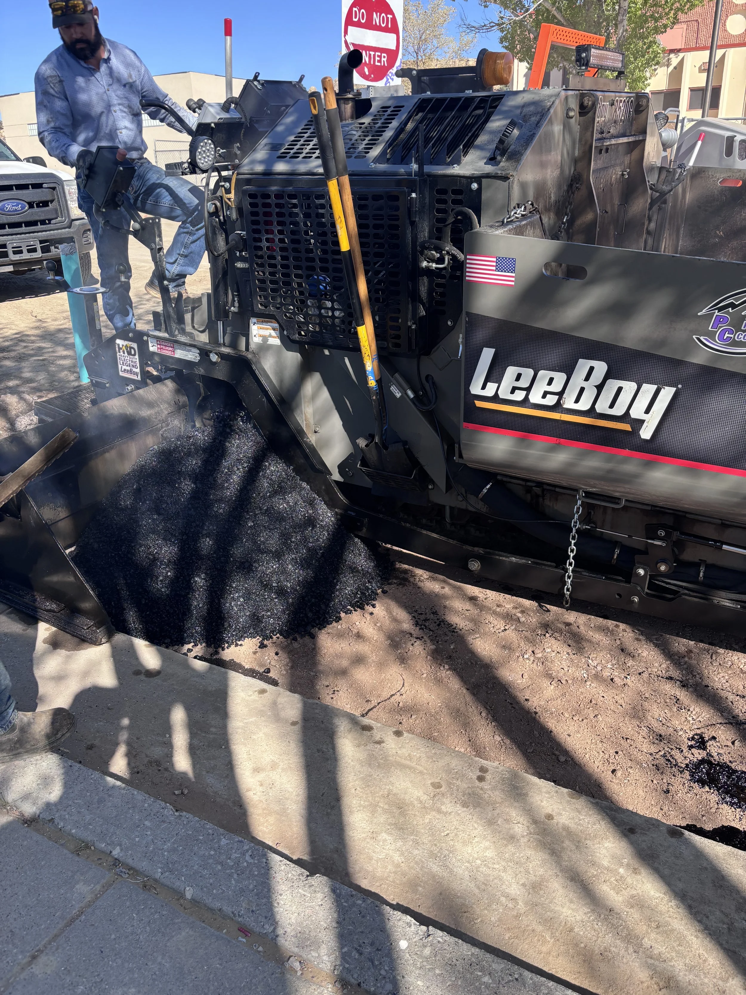 A worker in a blue shirt and baseball cap operates a black asphalt paver to lay asphalt on a sidewalk. The asphalt is black and fresh, with some spilled on the ground. There is a white Ford truck and a red 'Do Not Enter' sign in the background, along with trees and buildings.