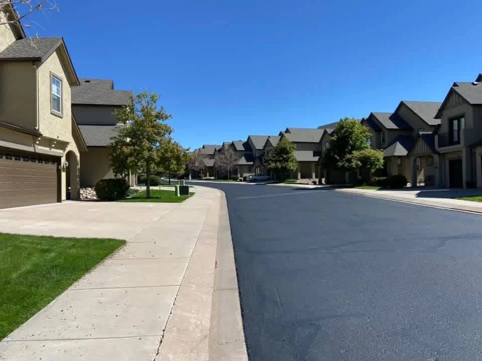 A quiet suburban neighborhood street lined with houses, trees, and well-maintained lawns under a clear blue sky.
