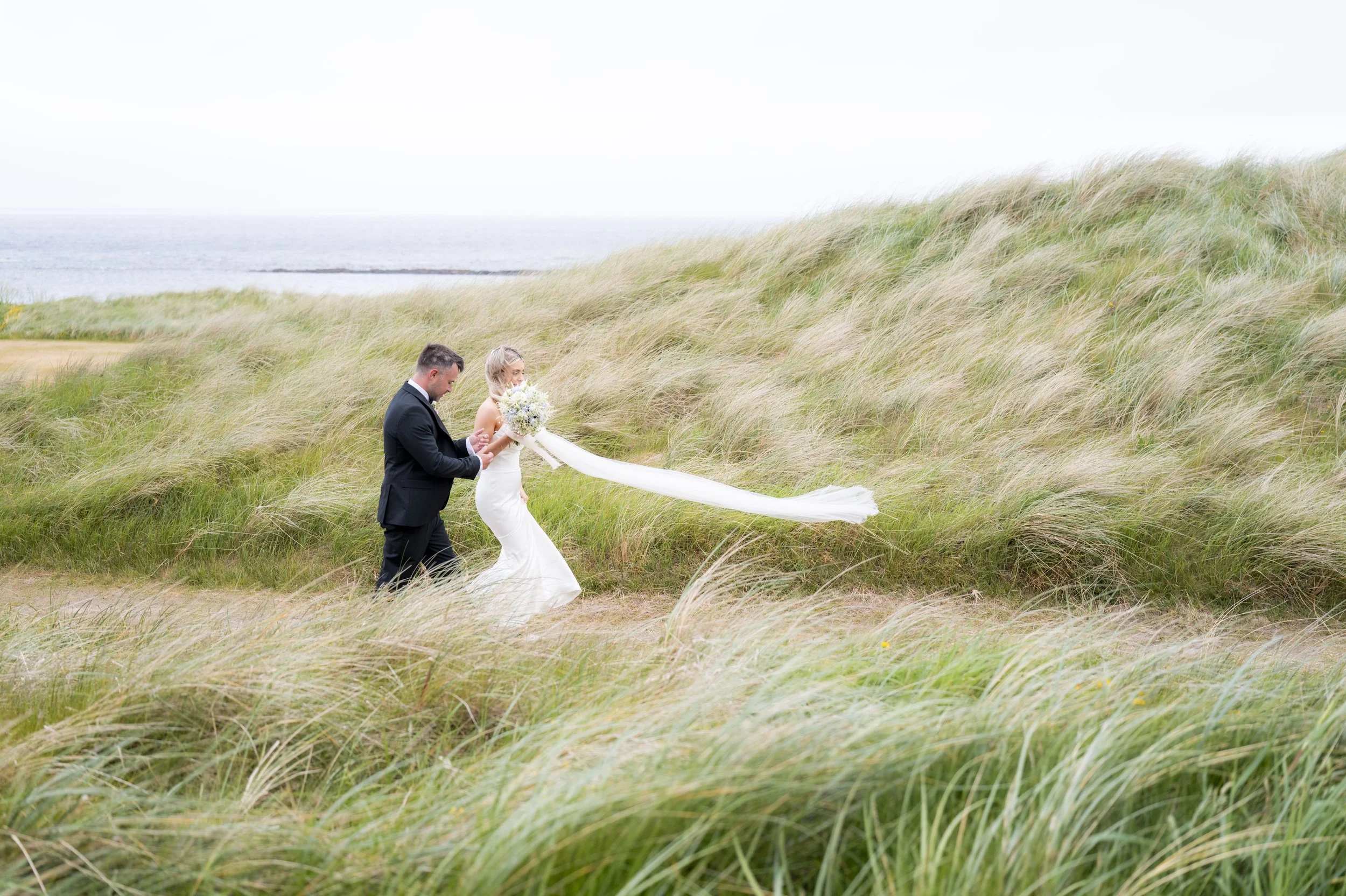 A bride and groom holding hands walking through tall grass on a beach with the ocean in the background.
