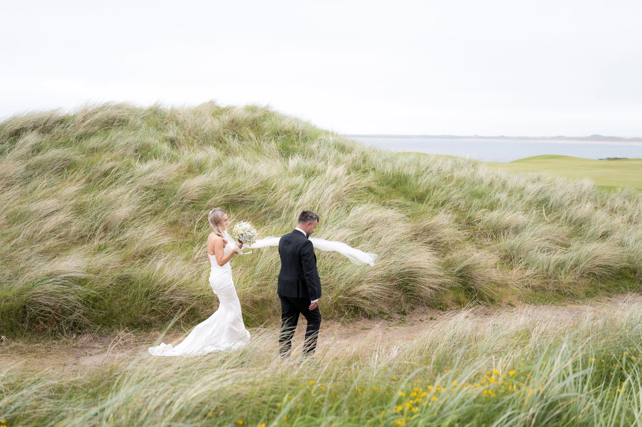 A bride and groom walking through tall grass near a body of water, with the bride holding a bouquet and wearing a dress with a long train, and the groom in a suit, during a wedding photo shoot.