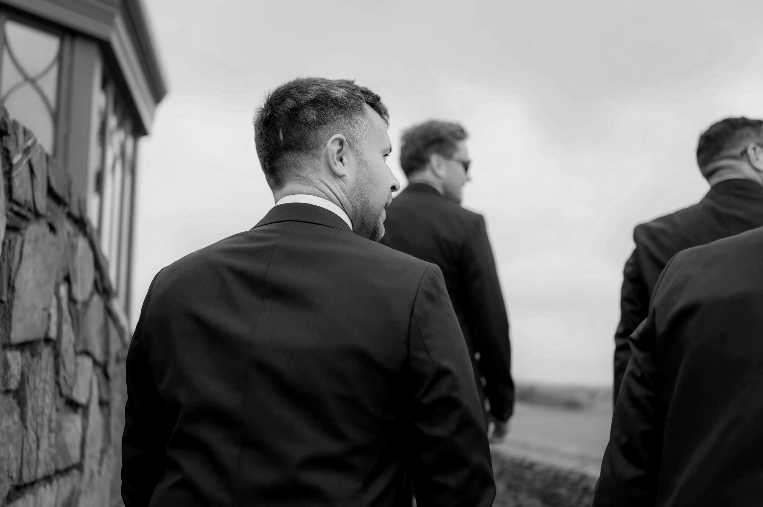 Black and white photo of men in suits sitting outdoors, facing away from the camera, with a stone wall on the left and open sky in the background.
