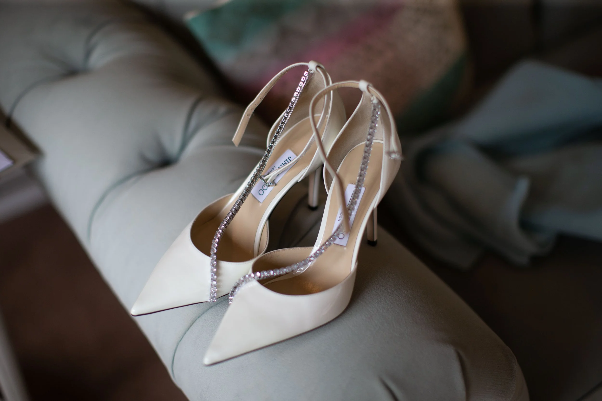 A pair of white high-heeled shoes with rhinestone straps resting on a light-colored cushioned surface.