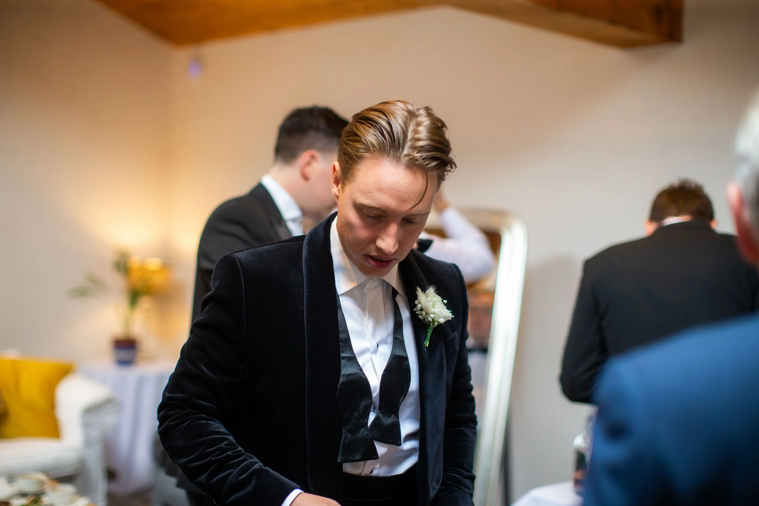 Young man in formal attire, possibly at a wedding, standing indoors, surrounded by other people in similar attire.