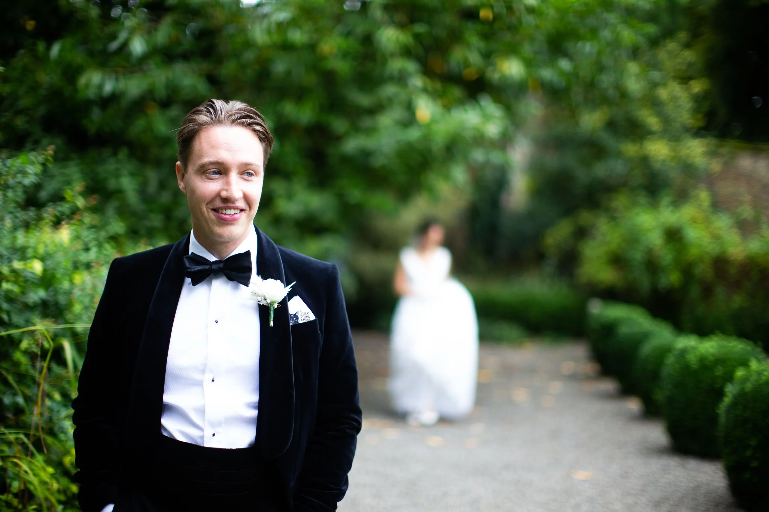A groom dressed in a black tuxedo with a white shirt and black bow tie, smiling and looking to the side outdoors, with a woman in a white dress blurred in the background surrounded by green foliage.