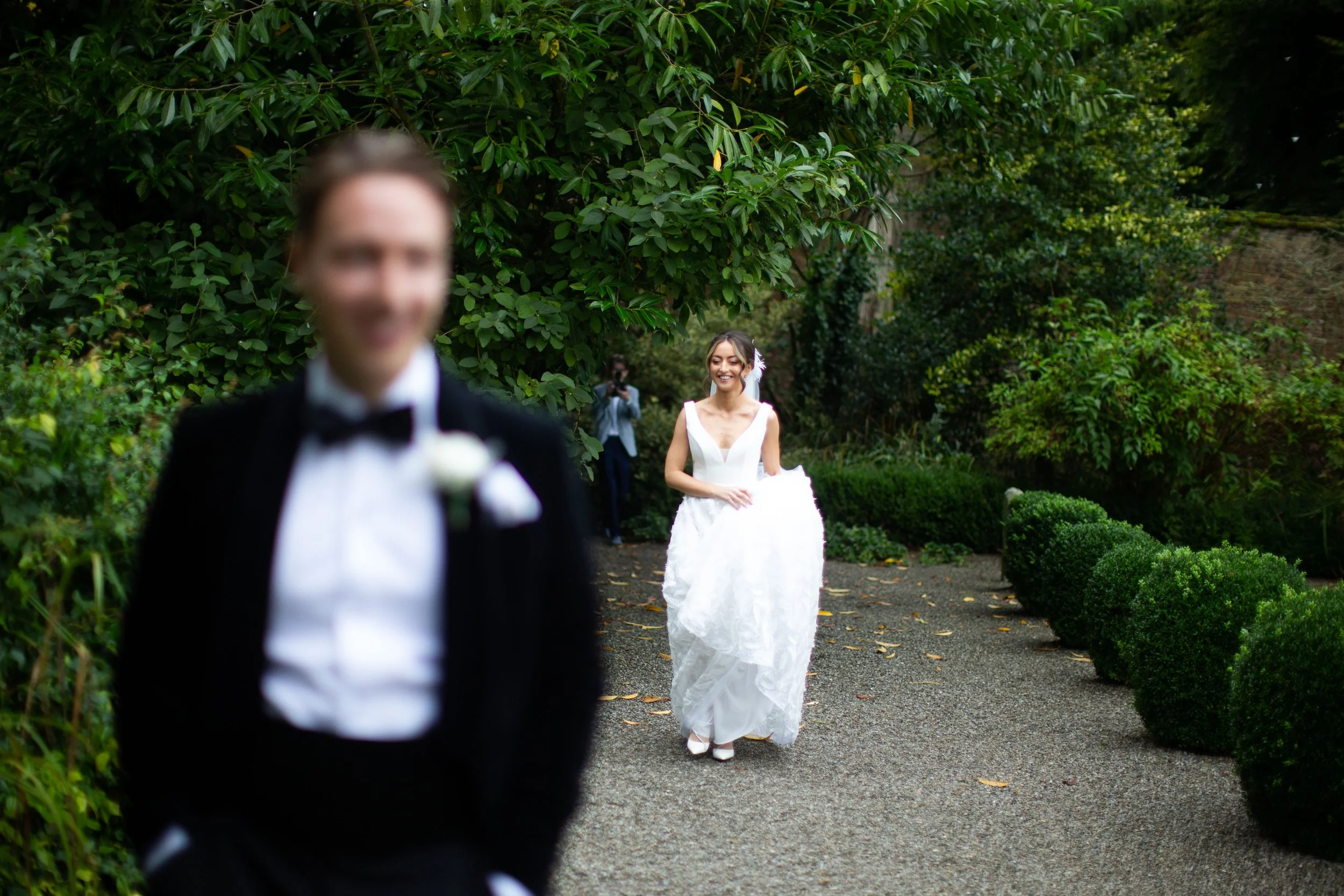 A bride smiling in a white wedding dress walking towards a groom in a black tuxedo for a wedding ceremony outdoors, with greenery in the background.