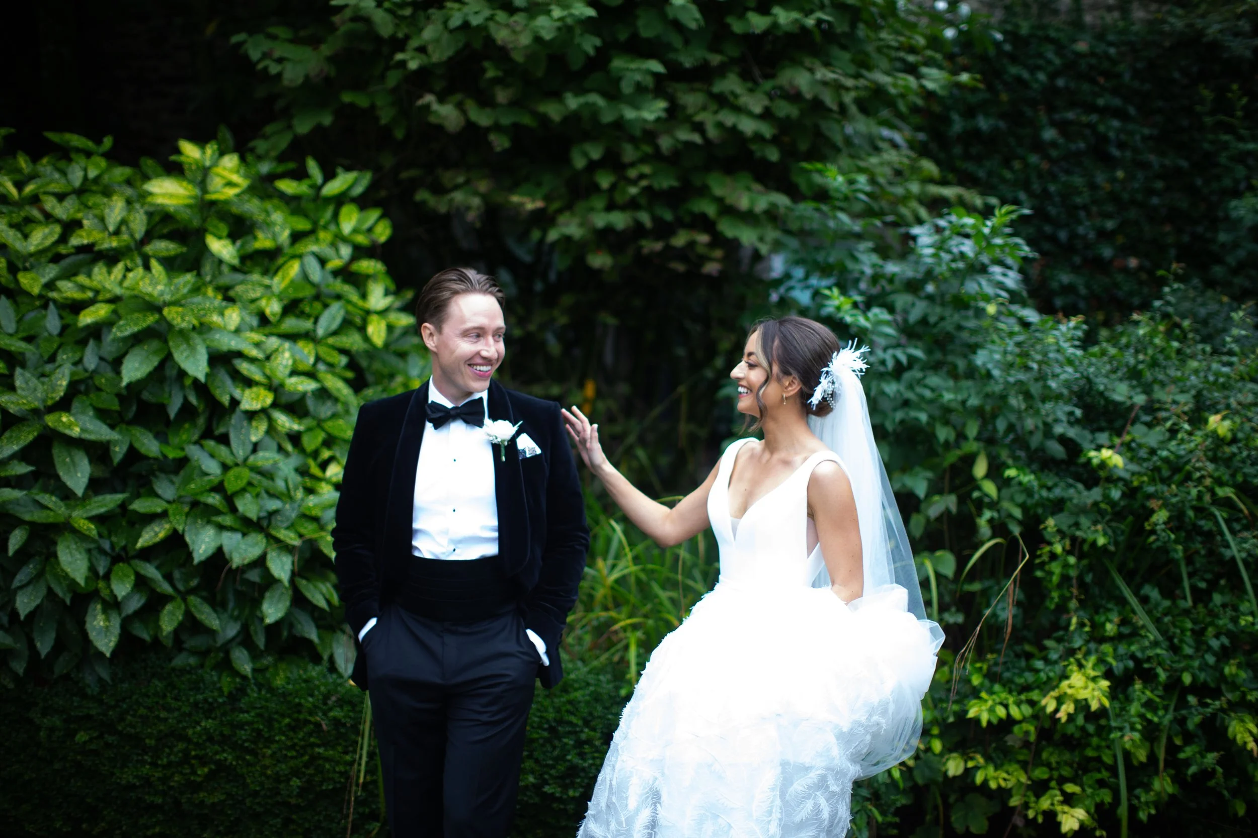 A bride and groom smiling and talking outdoors surrounded by green foliage, with the bride wearing a white wedding dress and veil, and the groom in a black tuxedo with a bow tie.