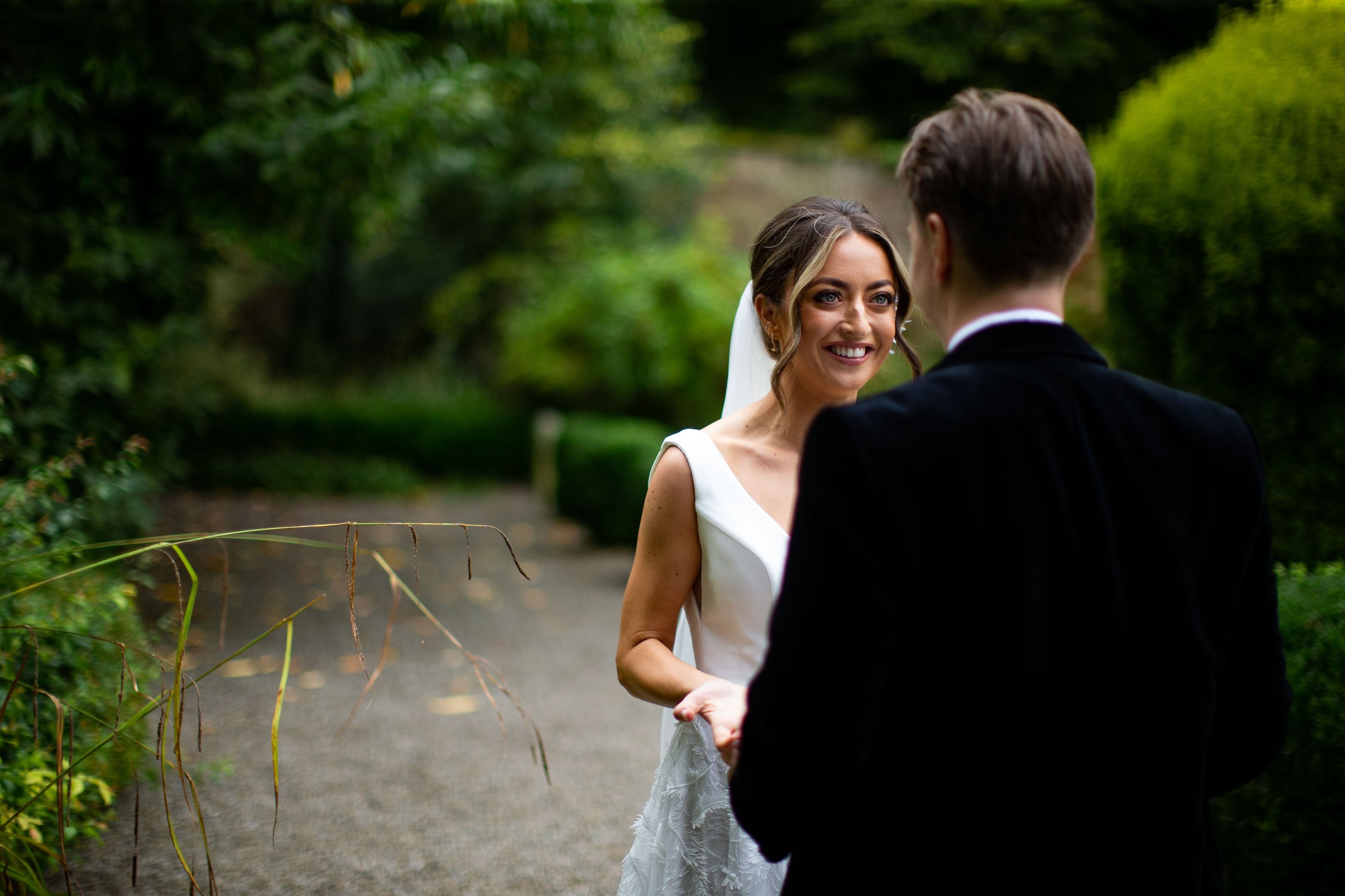 A bride and groom holding hands in a lush outdoor setting with green foliage, standing on a dirt path.